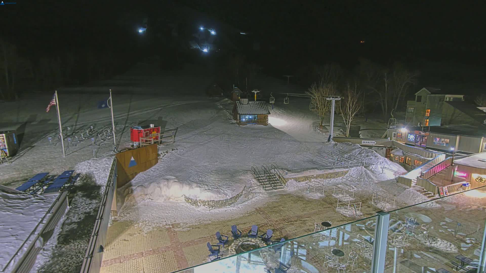 A snow-covered ski resort at night, illuminated by lights, showing a ski lift, buildings, and outdoor seating areas.