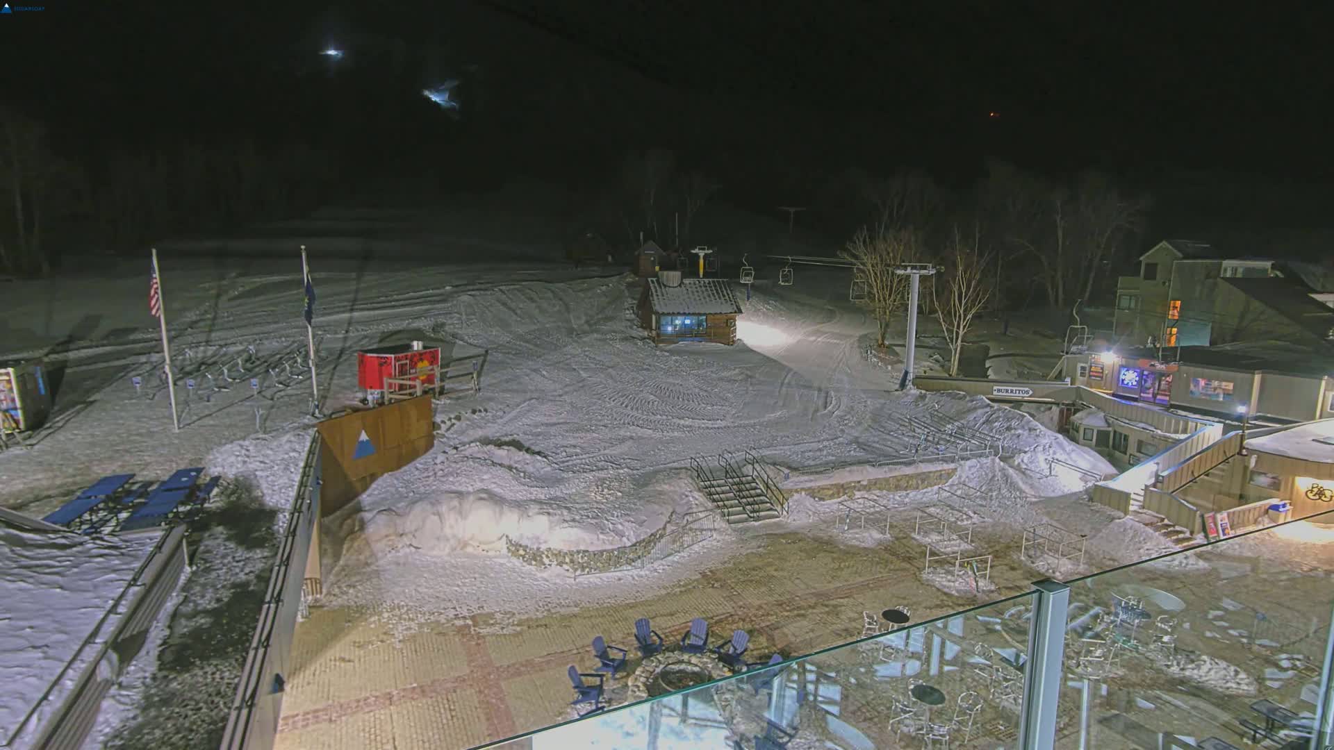 A snow-covered ski slope at night, illuminated by artificial lights, features a small building, ski lift, and outdoor seating area.
