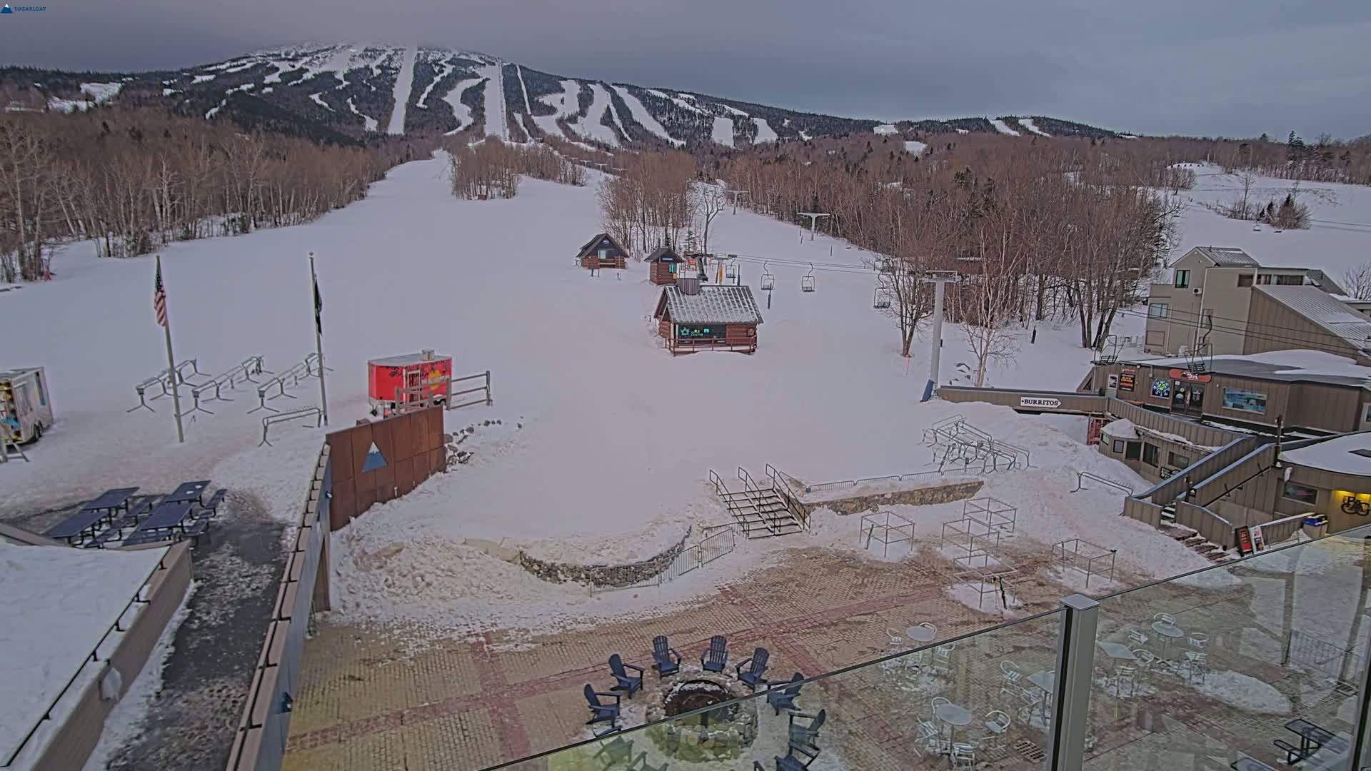 A snow-covered ski resort base area with several small buildings, a fire pit with chairs, and outdoor seating, overlooks a mountain with ski runs under an overcast sky.