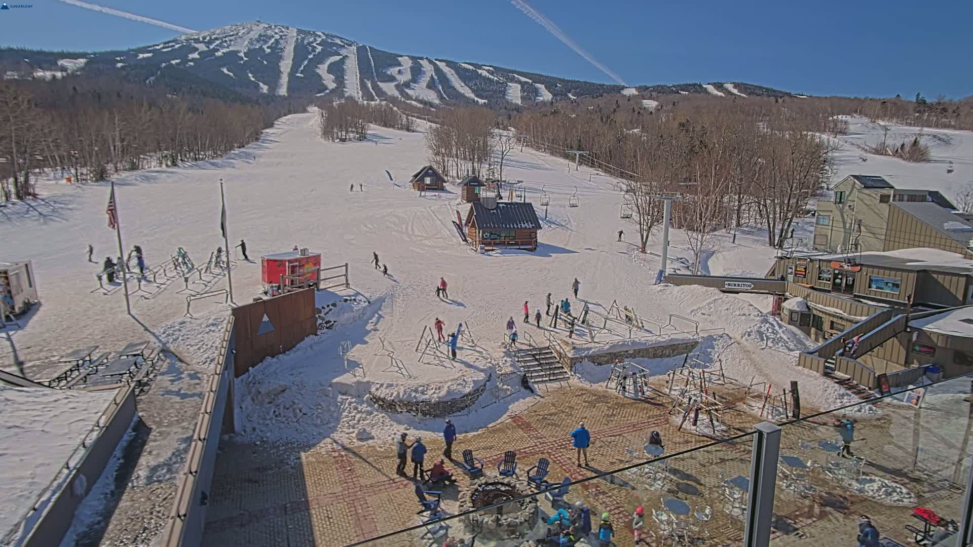 A sunny day at a snow-covered ski resort shows skiers and snowboarders on the slopes, with people gathered around a fire pit near a base lodge.