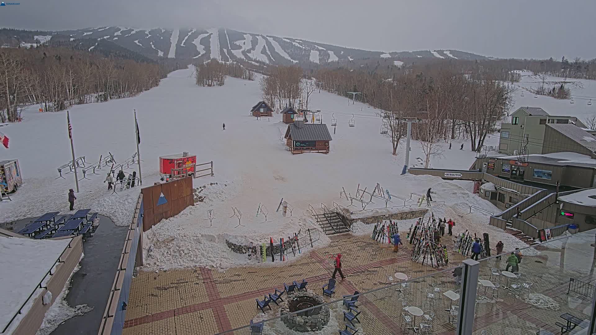 A snowy ski resort base area with several small buildings, ski racks, outdoor seating, and a fire pit, under an overcast sky.