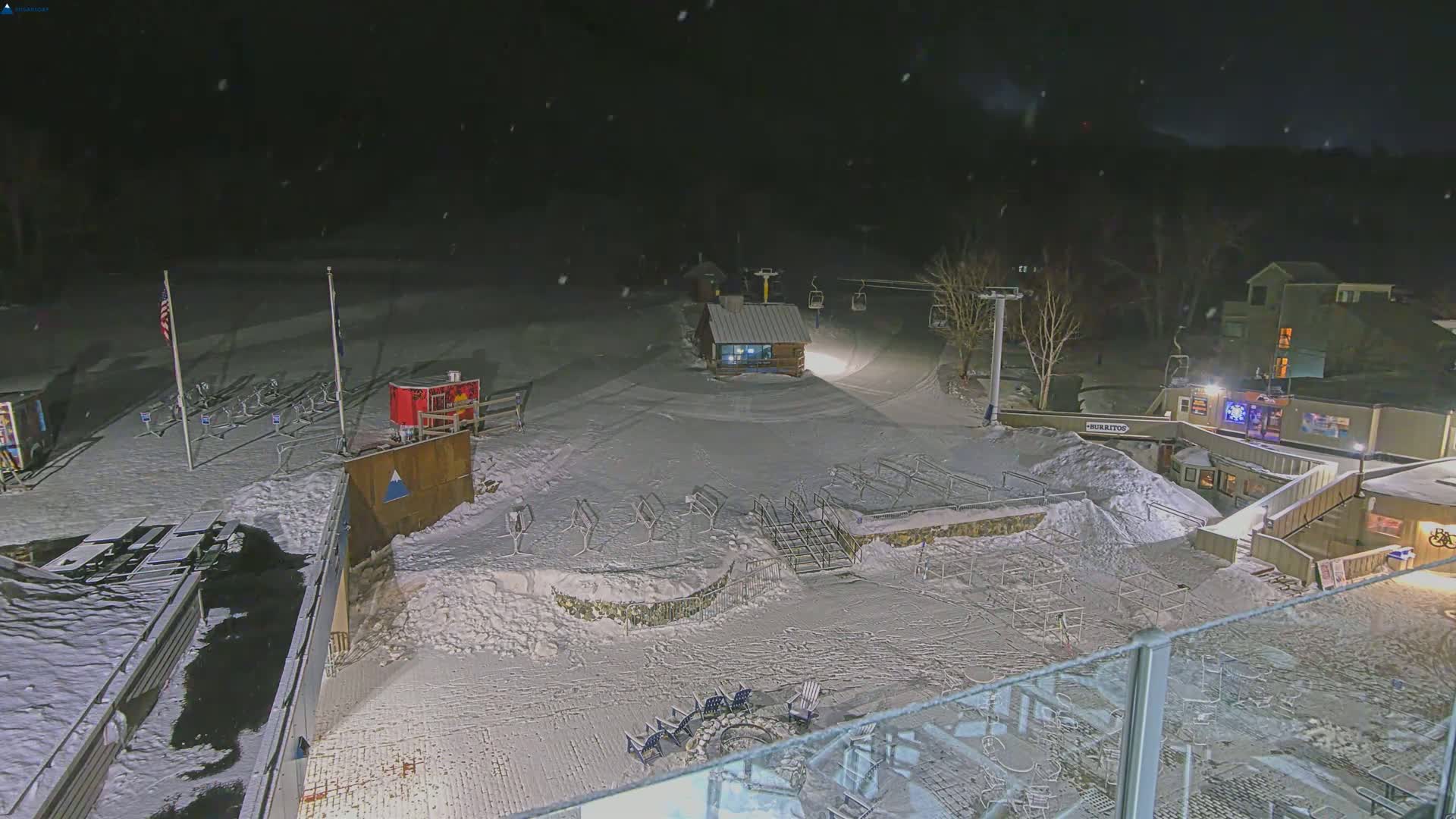 A snow-covered ski resort at night, lightly snowing, with a chairlift, a small building, and outdoor seating areas.