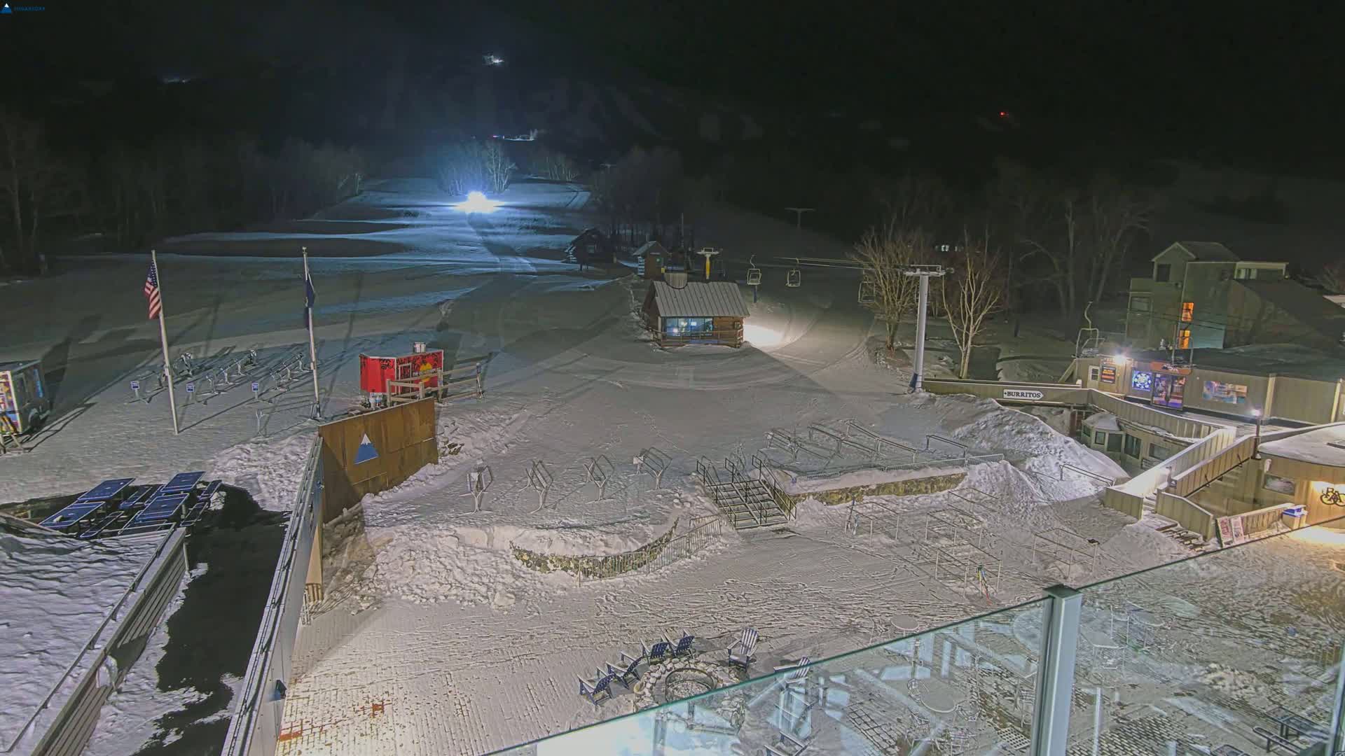 A snow-covered ski resort at night, illuminated by artificial lights, with a ski lift and several buildings visible.