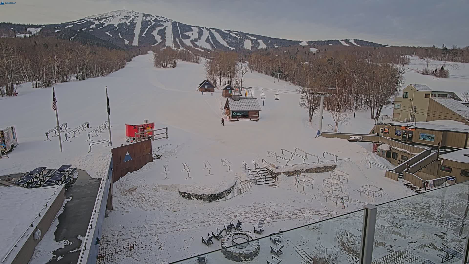 A snow-covered ski resort base area, featuring several small buildings, ski lifts, and picnic tables, sits below a large snow-covered mountain under a cloudy sky.