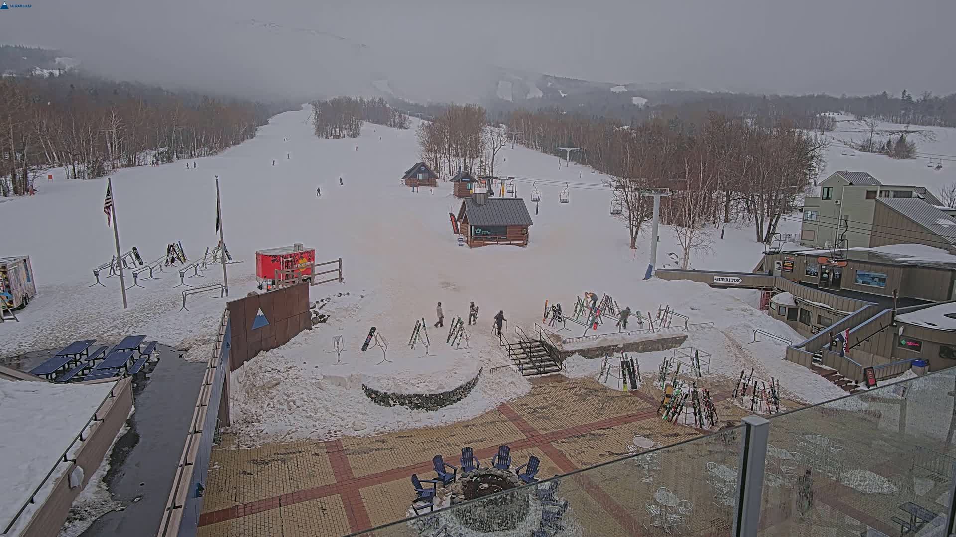 A snowy ski resort base area on an overcast day, showing skiers, ski racks, small buildings, and outdoor seating around a fire pit.