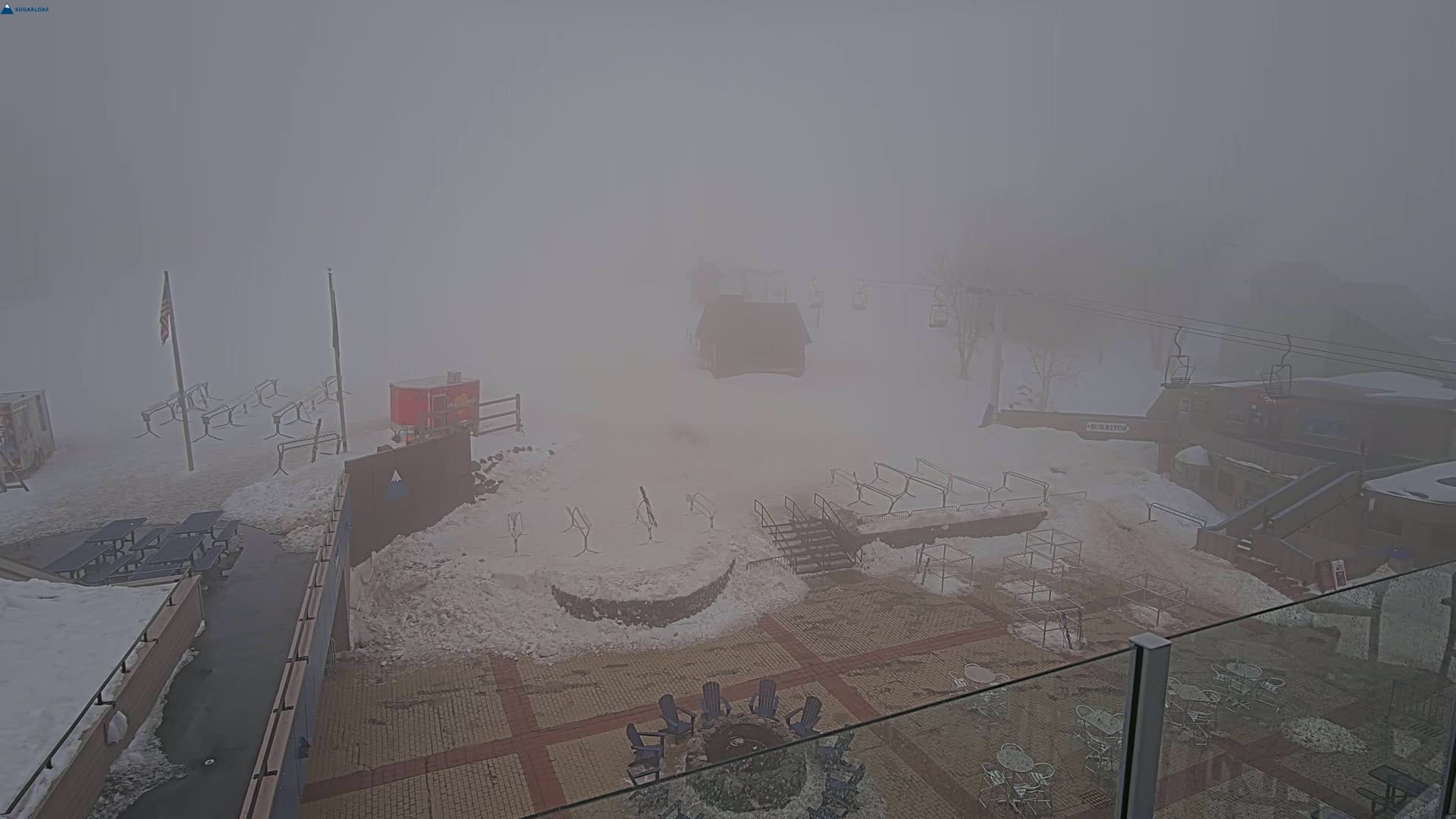 A snow-covered ski resort is shrouded in thick fog, with several buildings, chairs, and ski lifts barely visible.