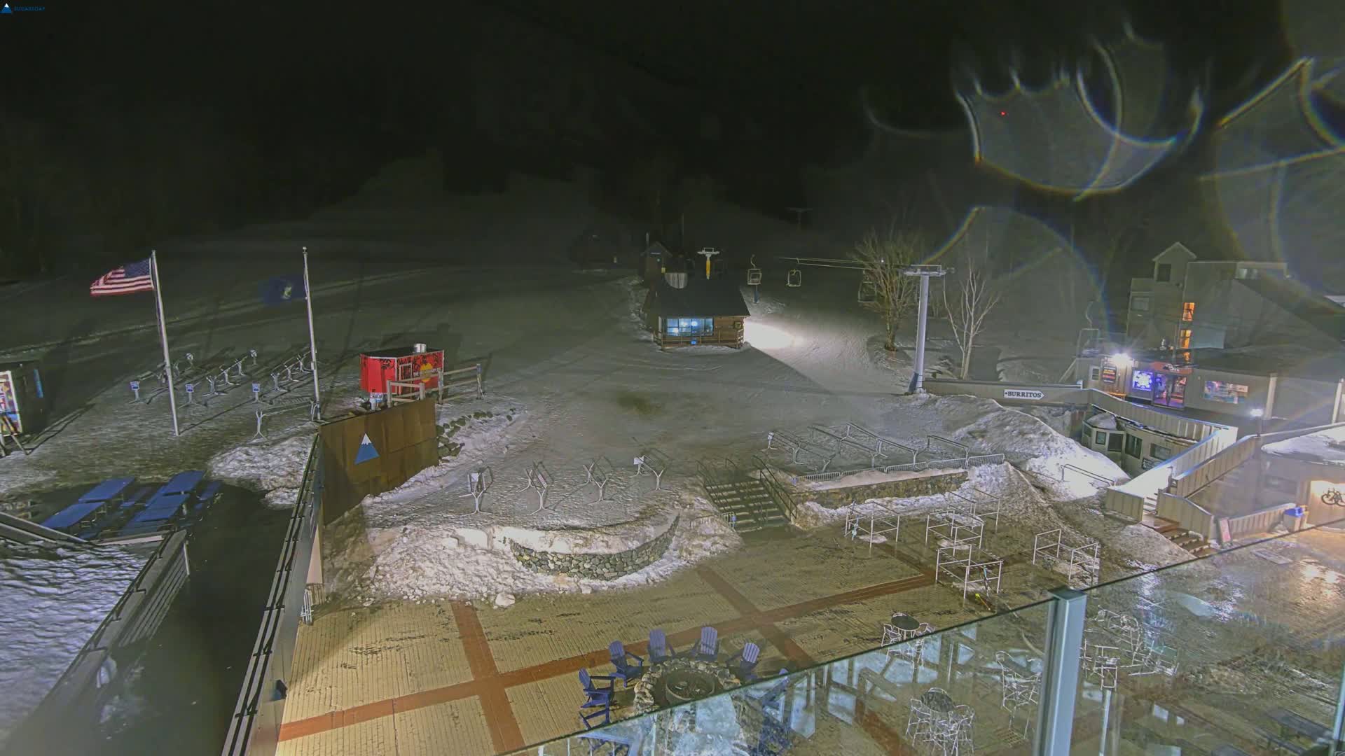 A nighttime, snow-covered ski resort base area with outdoor seating, a small building, and a ski lift visible in the background.