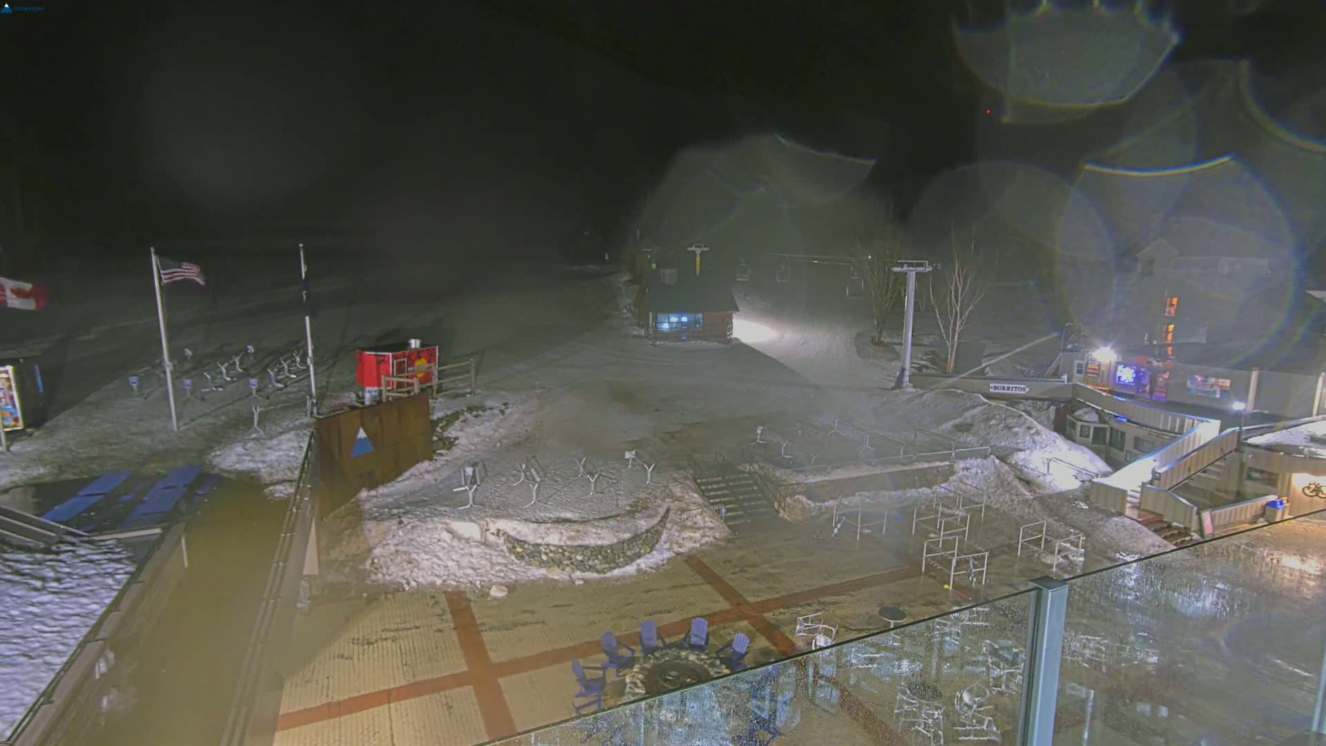 A nighttime view of a snow-covered ski resort base area with a patio featuring outdoor seating, under a slightly hazy sky with rain or snow.