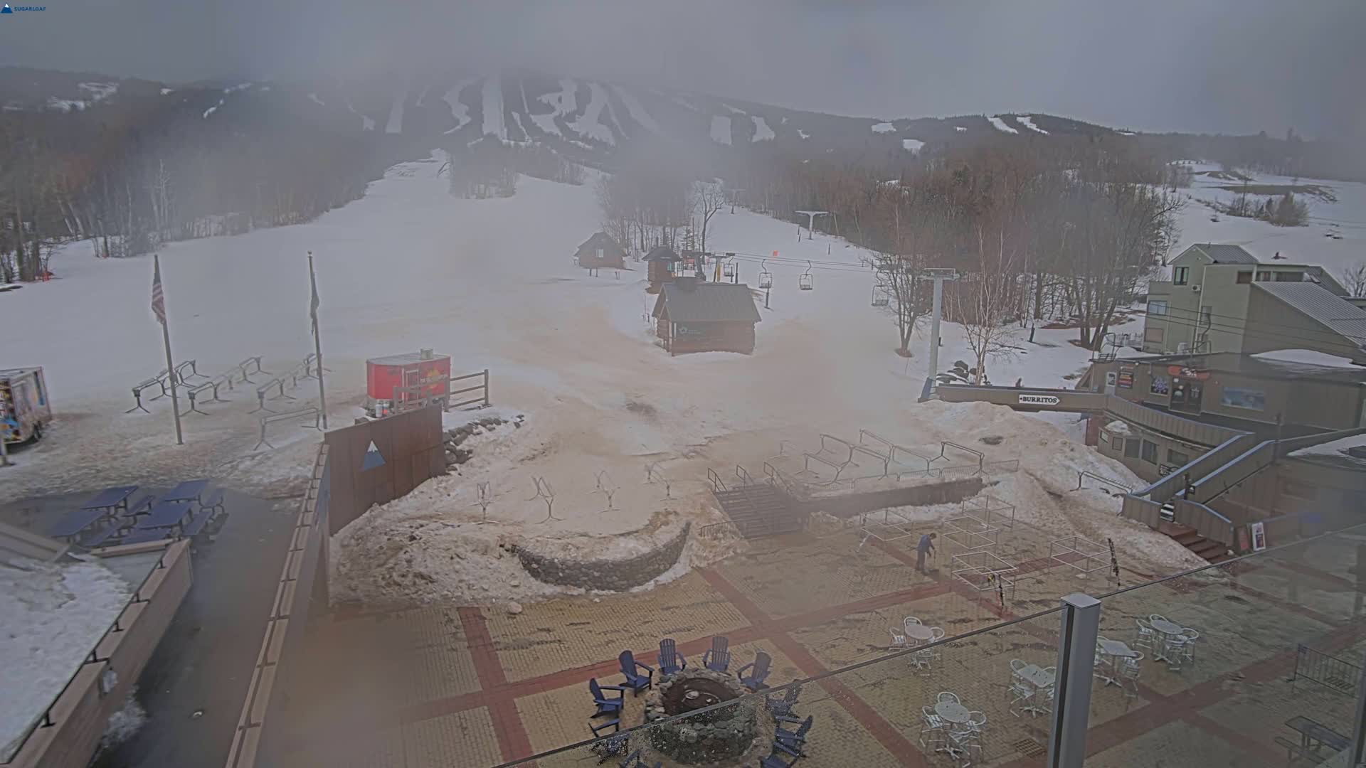 A foggy view of a snow-covered ski resort base area with several buildings, a fire pit, and outdoor seating.