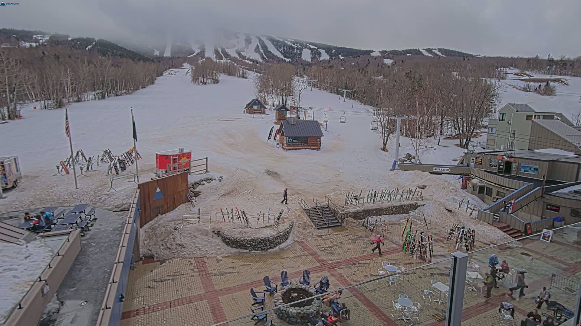 A snow-covered ski resort base area with several buildings, ski racks, and people walking around on an overcast day.