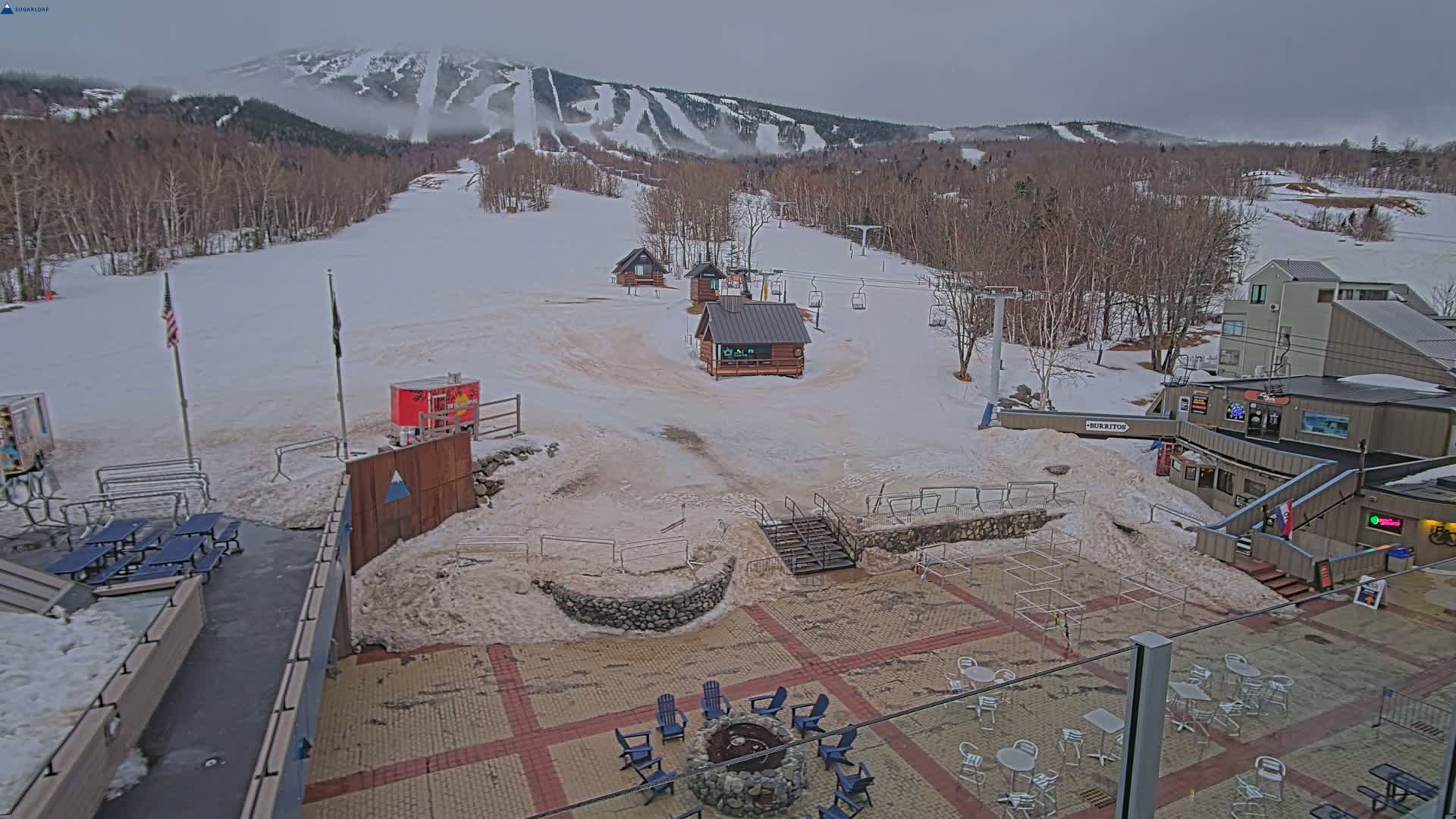 A snow-covered ski resort base area with several buildings, a fire pit, and outdoor seating, under an overcast sky.
