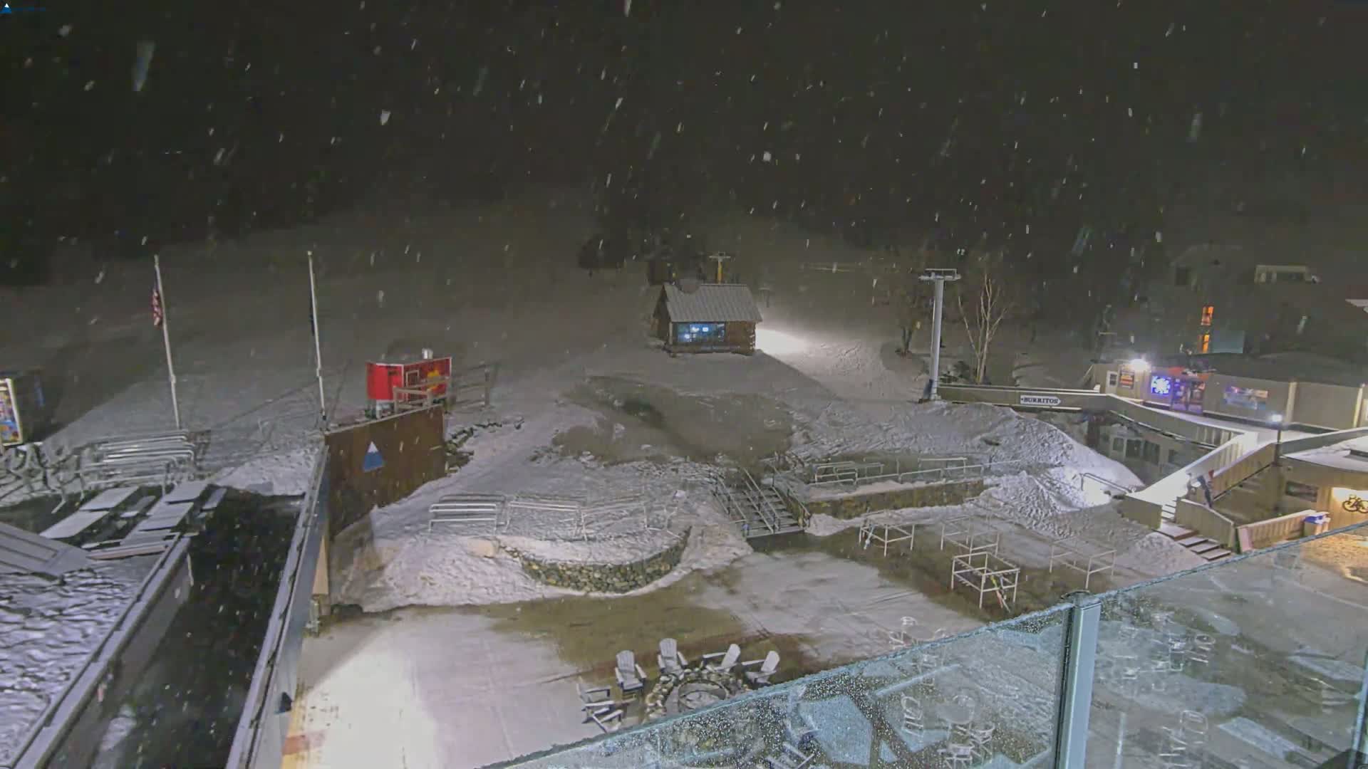 A nighttime snow scene at a ski resort shows snow-covered ground, buildings, and outdoor seating areas with a fire pit, under falling snow.