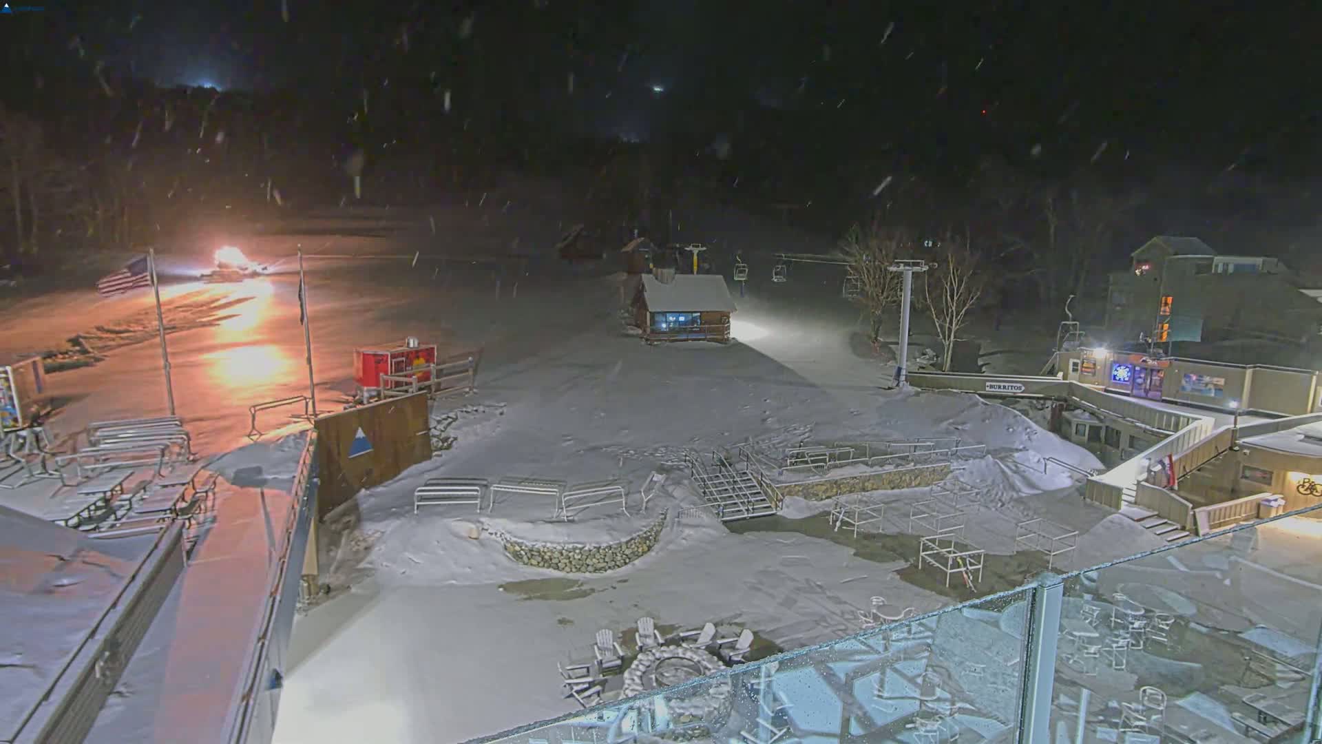 A snow-covered ski resort at night, lightly snowing, with a fire pit and several buildings illuminated.