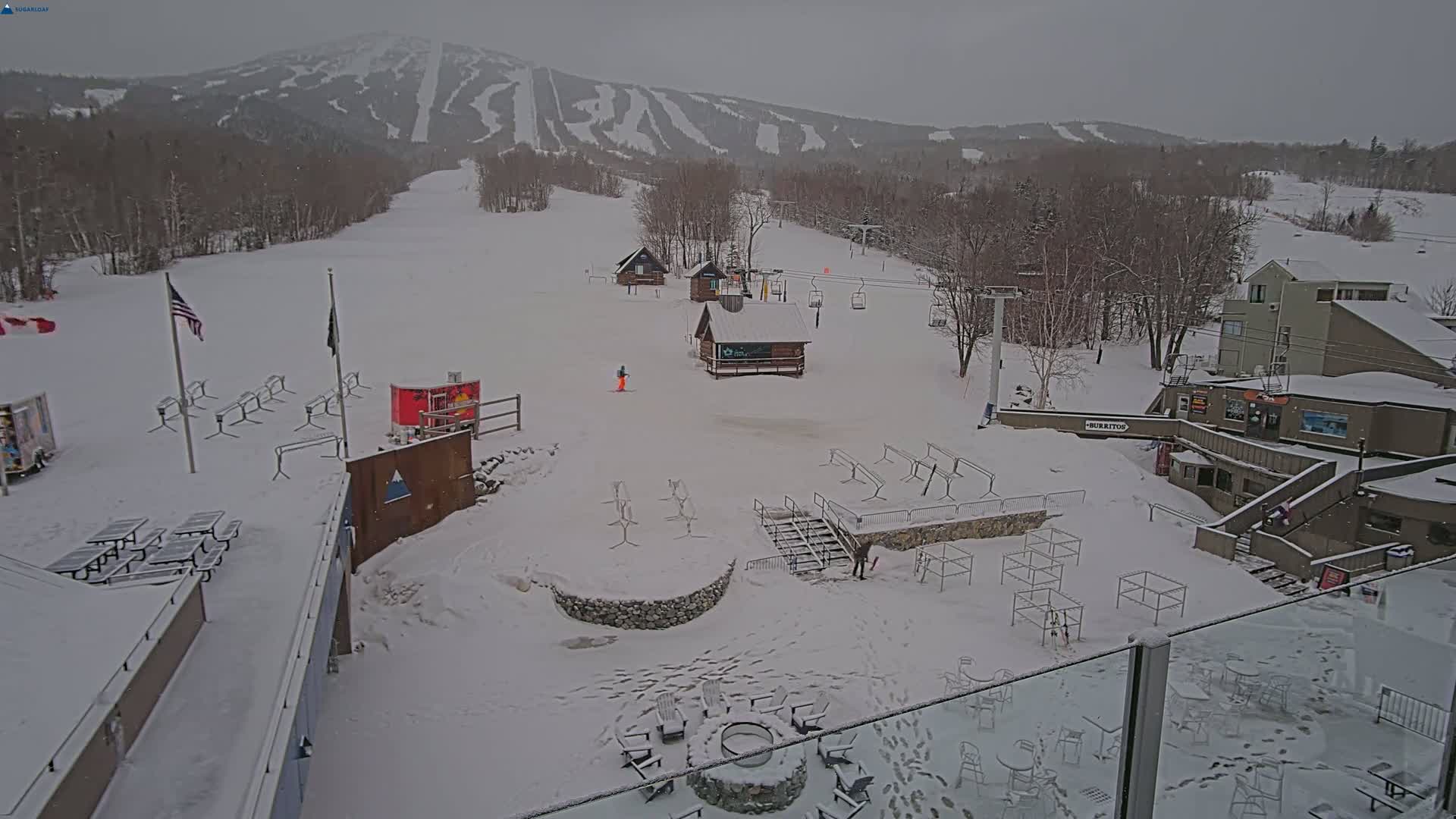 A snow-covered ski resort base area with several buildings, picnic tables, ski racks, and a chairlift visible in the background under an overcast sky.