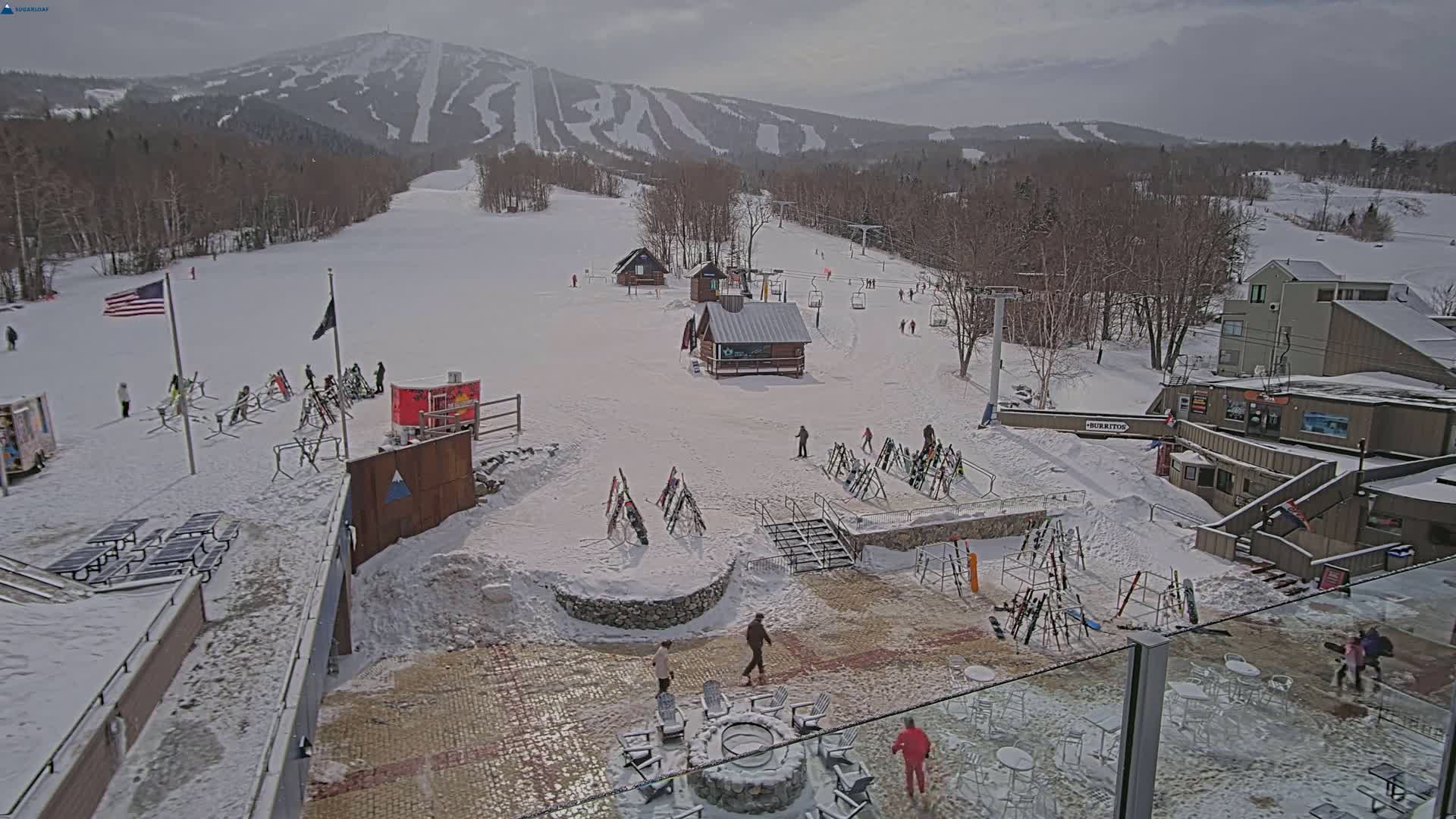 A snowy ski resort base area with several small buildings, ski racks, and people, overlooking a mountain with ski runs under an overcast sky.