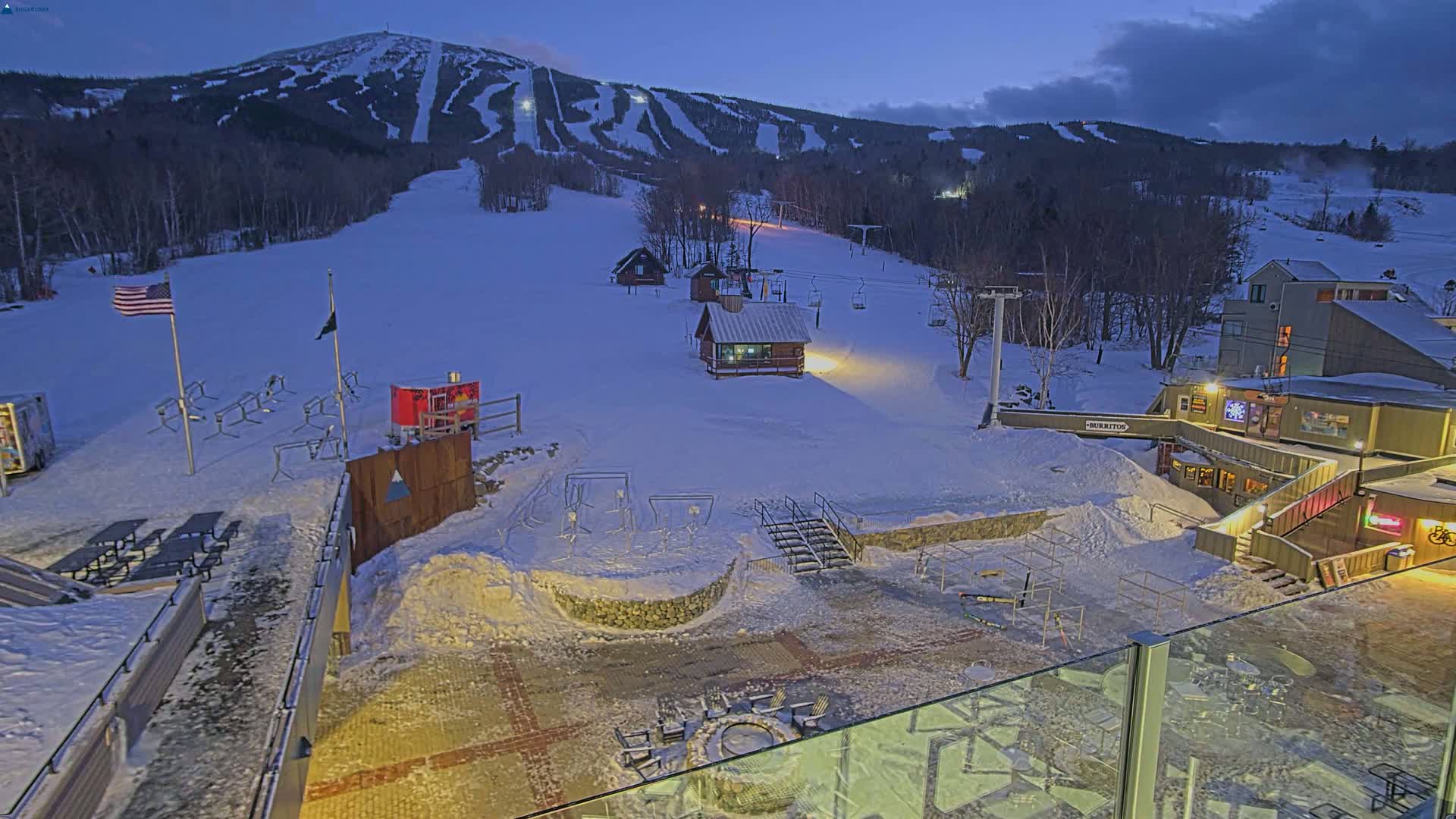 A snow-covered ski resort at twilight, with a mountain in the background, features several buildings, ski lifts, and outdoor seating areas around a fire pit.