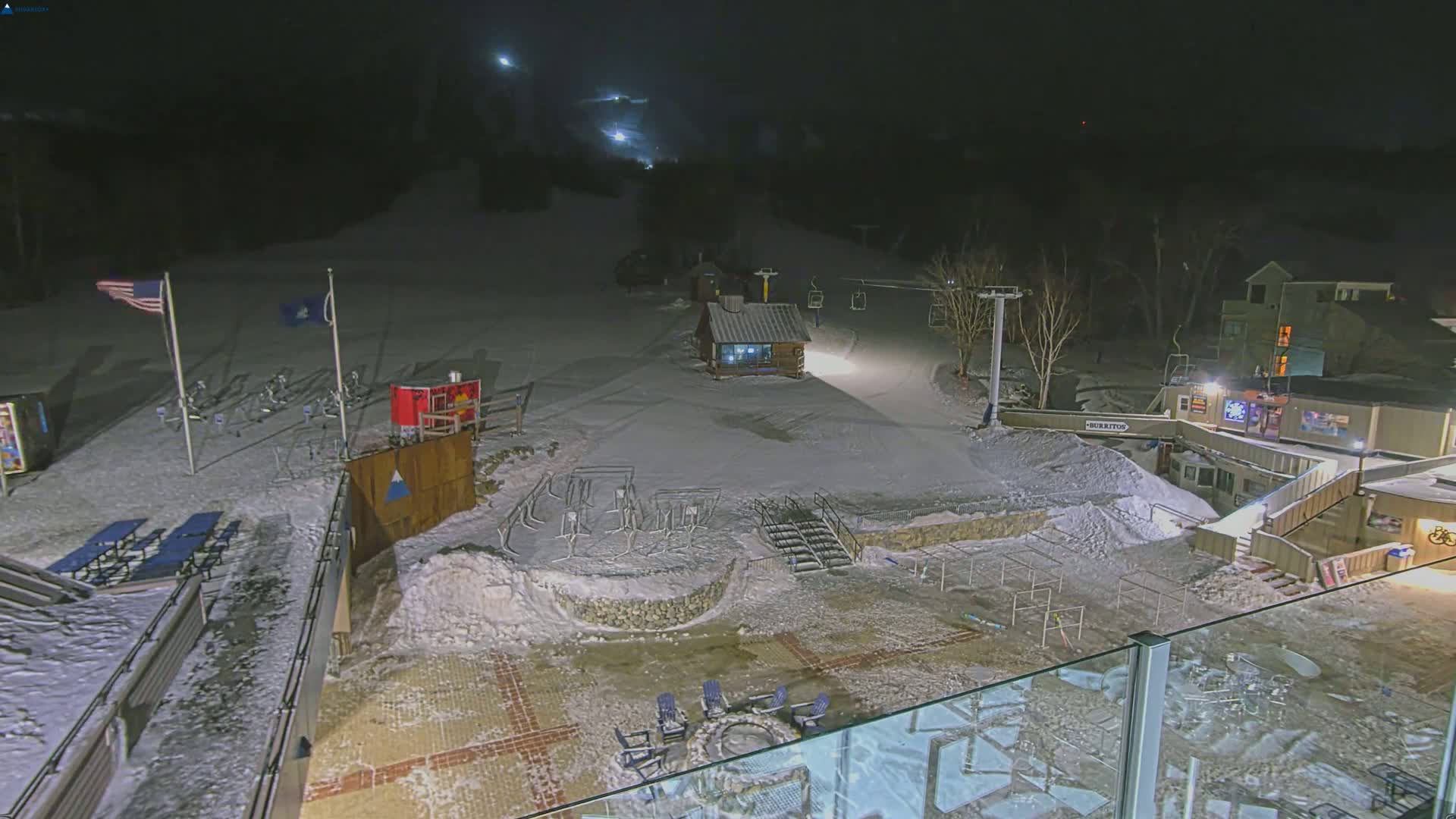 A snow-covered ski resort at night, illuminated by artificial lights, shows a ski lift, a small building, and outdoor seating areas.