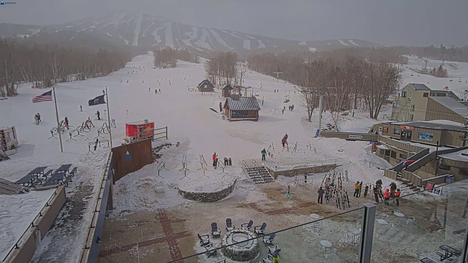 A snowy ski resort with several skiers and snowboarders visible on the slopes, a few buildings, and a fire pit area in the foreground under an overcast sky.