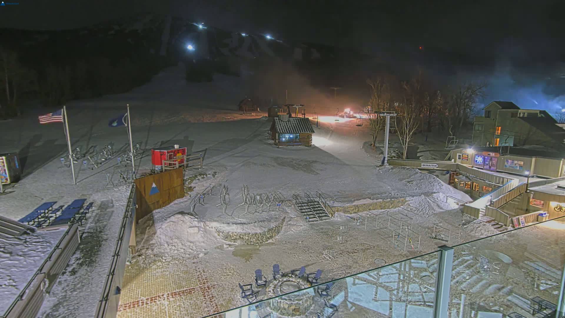 A snow-covered ski resort at night, illuminated by lights, with several buildings and a lift visible.