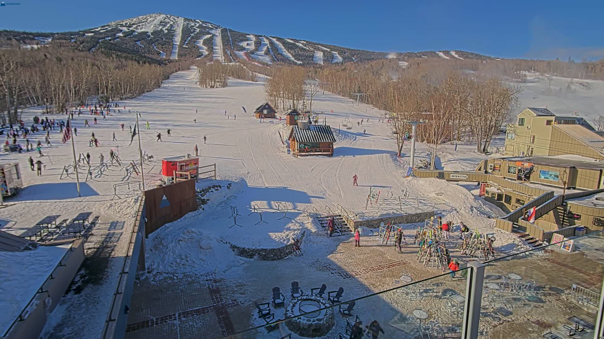 A sunny day at a snow-covered ski resort shows skiers and snowboarders on the slopes, with buildings and a fire pit visible in the base area.