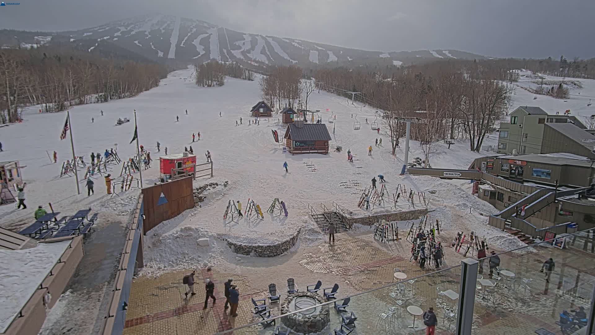 A snowy ski resort with several skiers and snowboarders on the slopes, ski racks, buildings, and outdoor seating areas under a mostly cloudy sky.