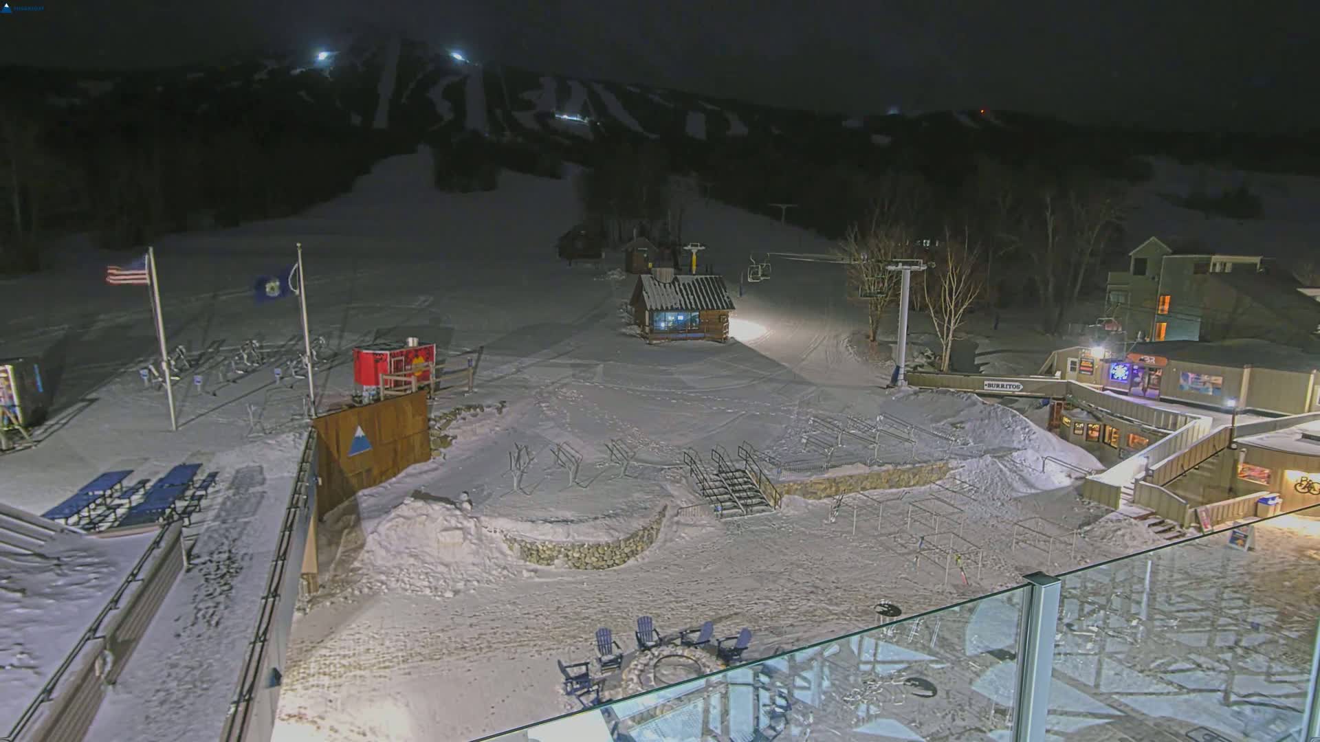 A snow-covered ski resort at night, illuminated by artificial lights, with several buildings and ski lifts visible.