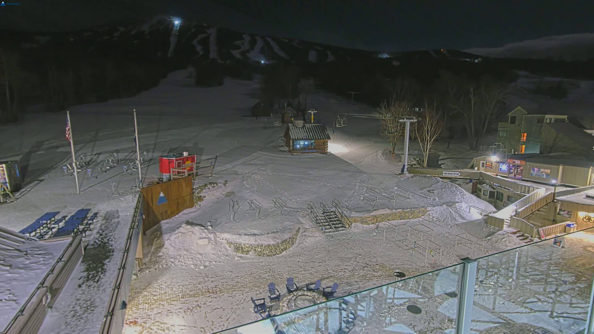 A snow-covered ski resort at night, illuminated by artificial lights, showing a ski lift, small buildings, and outdoor seating areas.
