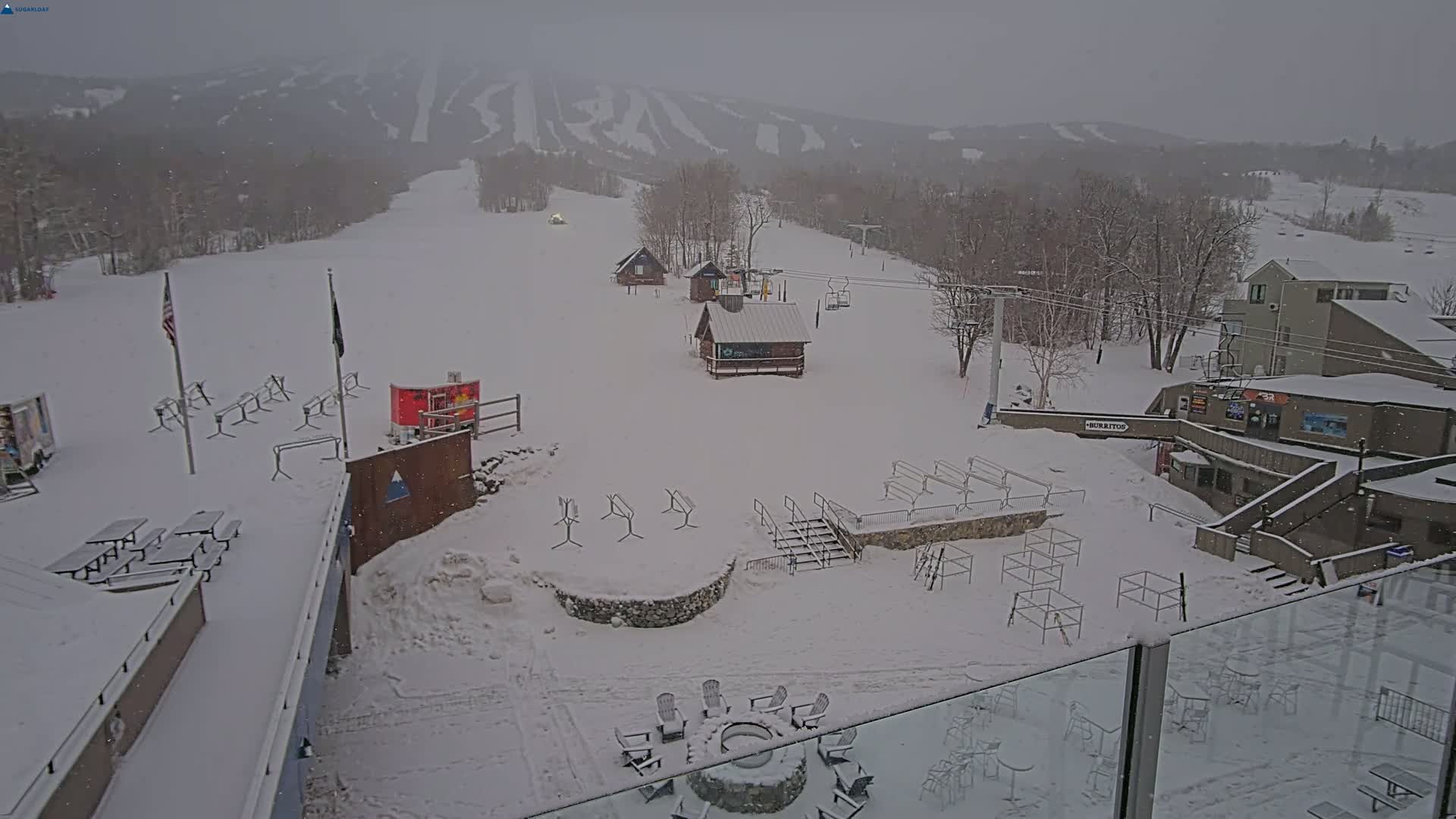 A snow-covered ski resort base area with several buildings, outdoor seating, and a ski lift in the background during a light snowfall.