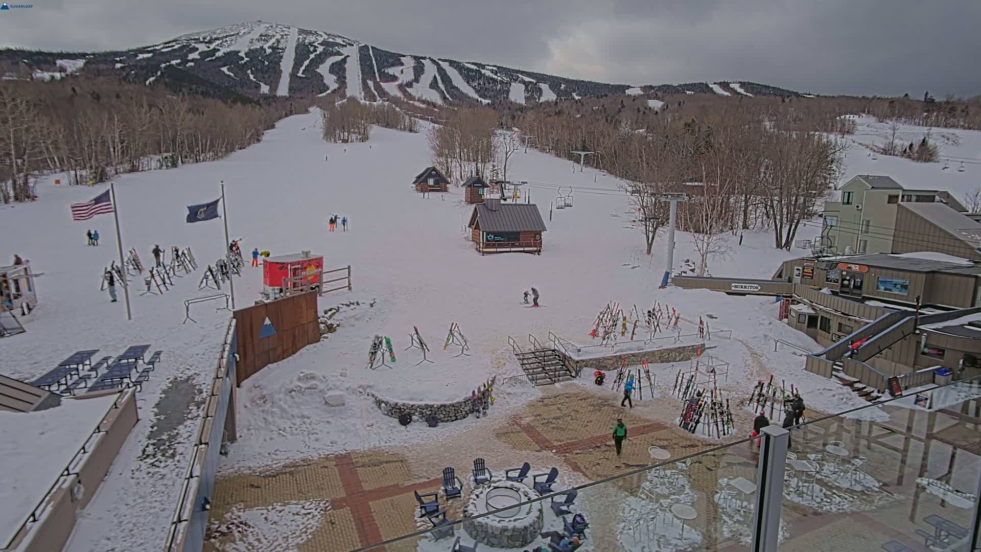 A snowy ski resort base area with several buildings, ski racks, and people, overlooks a mountain with ski trails under an overcast sky.