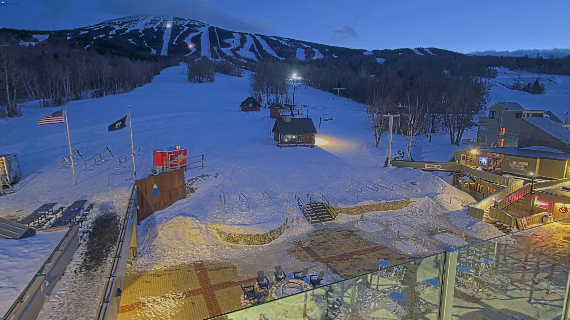 A snow-covered ski resort at dusk, with a mountain in the background, features several buildings, ski lifts, and outdoor seating areas.