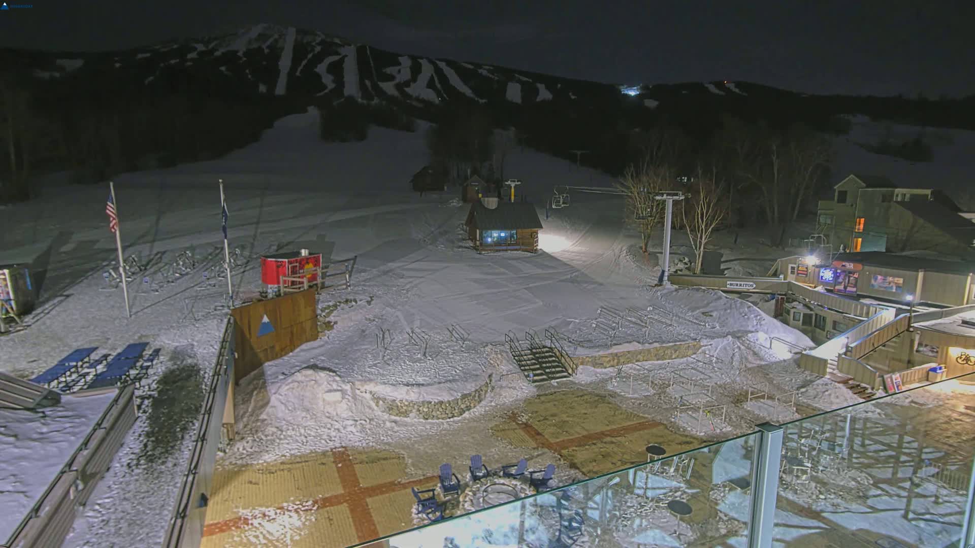 A snow-covered ski resort at night, illuminated by artificial lights, shows a small building, ski lifts, and outdoor seating areas.