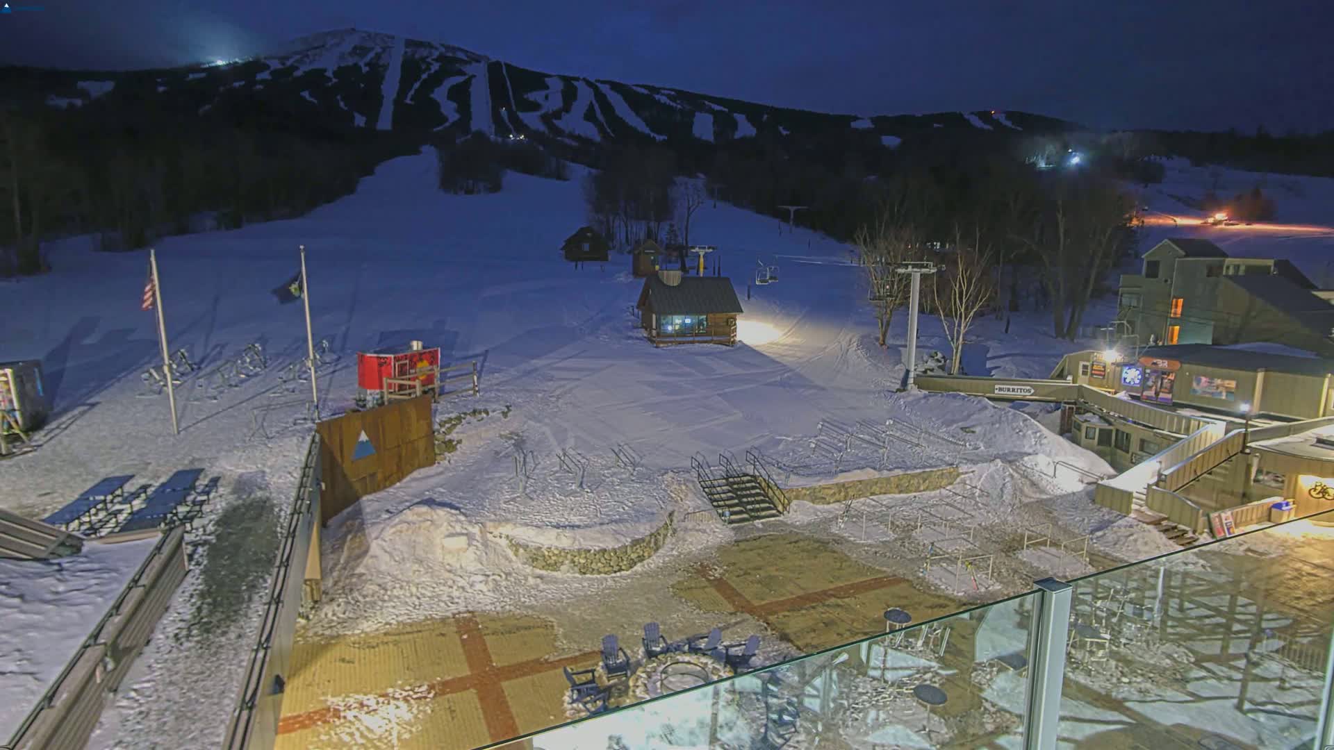 A snow-covered ski resort at night, illuminated by artificial lights, shows a base area with buildings, ski lifts, and a fire pit.