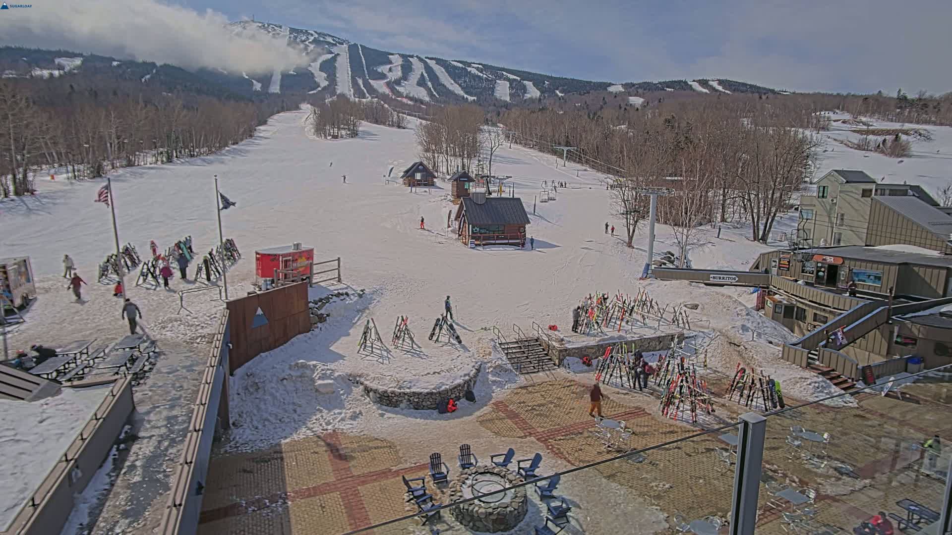 A snow-covered ski resort base area with people, ski racks, and small buildings, under a partly cloudy sky.