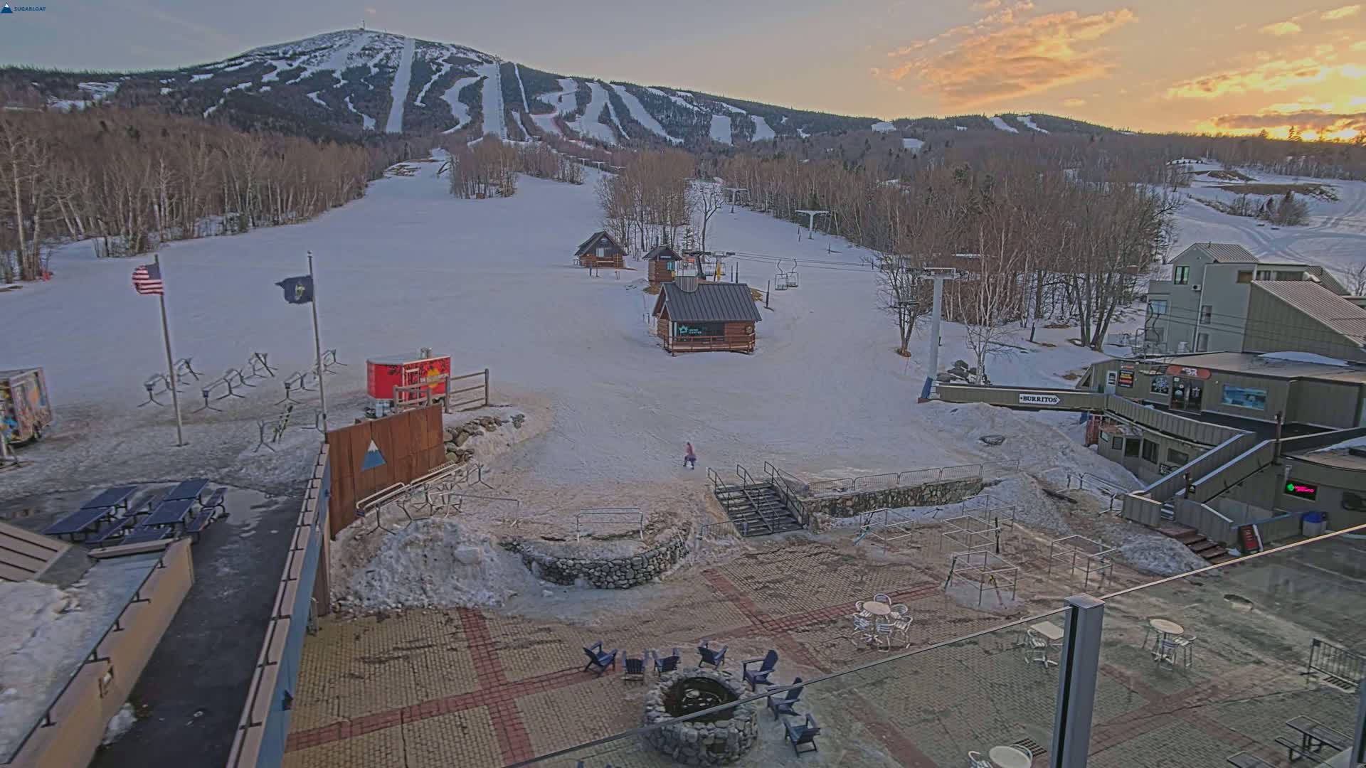 A snow-covered ski resort at sunset, featuring several small buildings, ski lifts, and outdoor seating areas.