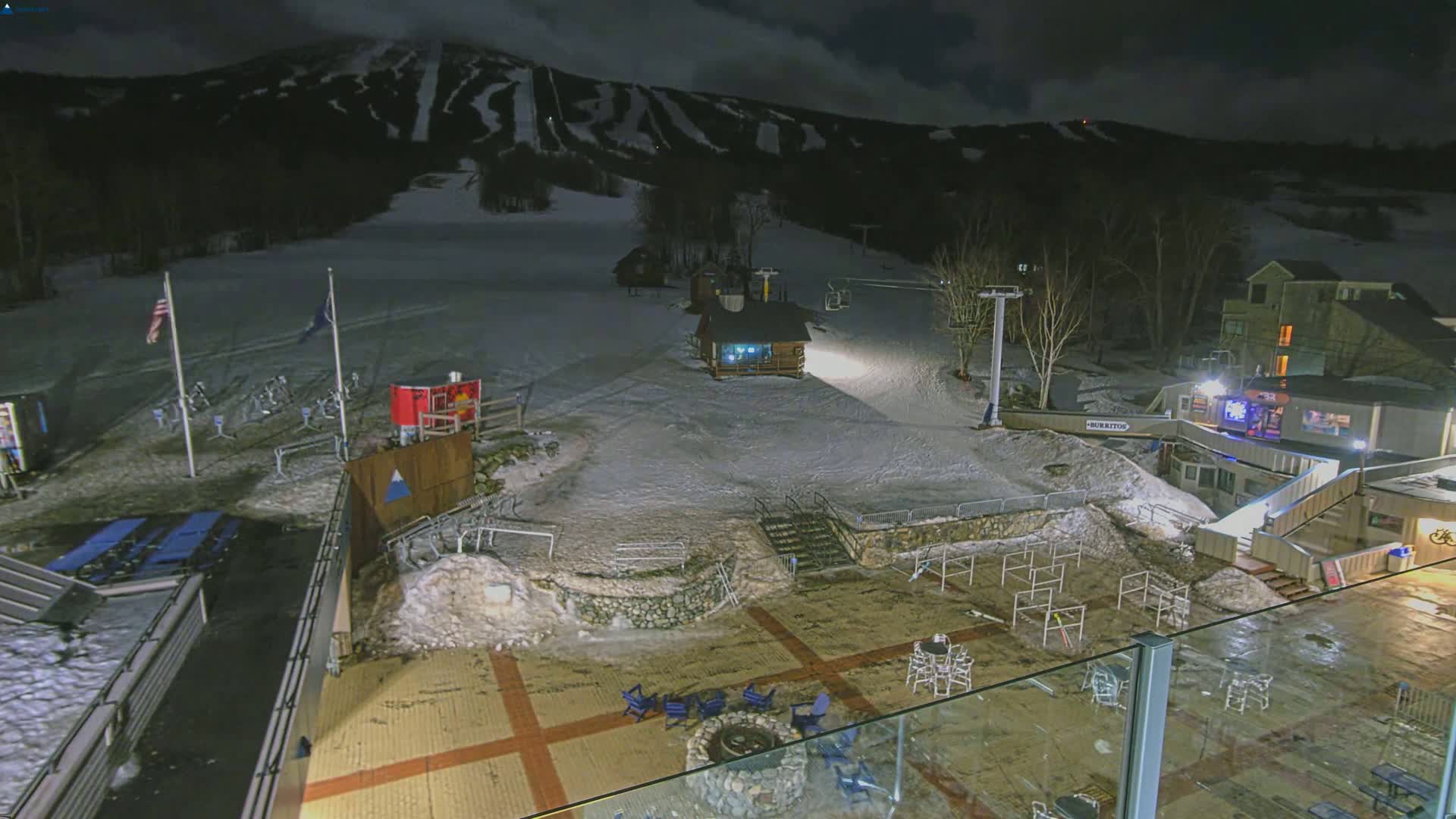 A snow-covered ski resort at night, illuminated by artificial light, features a base area with seating, a fire pit, and a ski lift against a backdrop of a snow-dusted mountain.