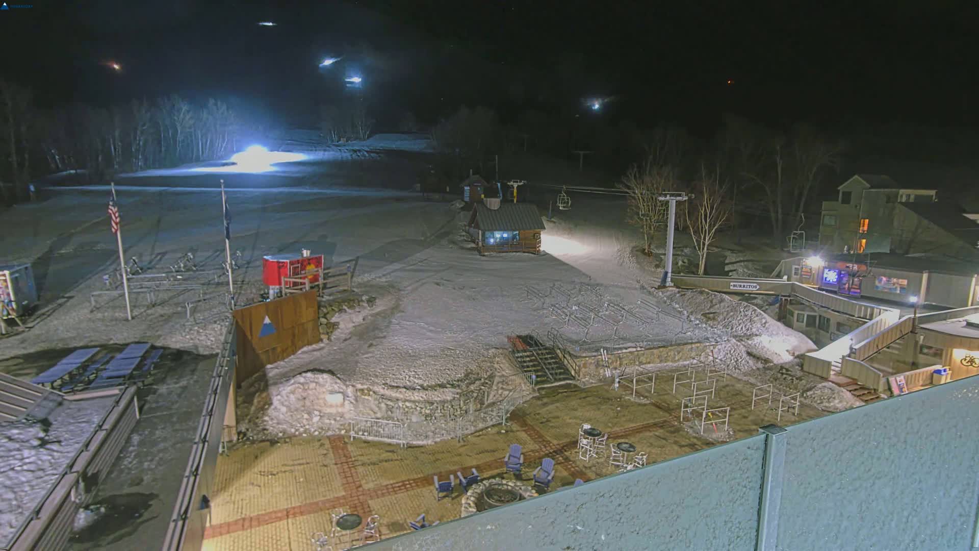 A snow-covered ski resort at night, illuminated by artificial lights, shows a ski lift, small buildings, and outdoor seating areas.