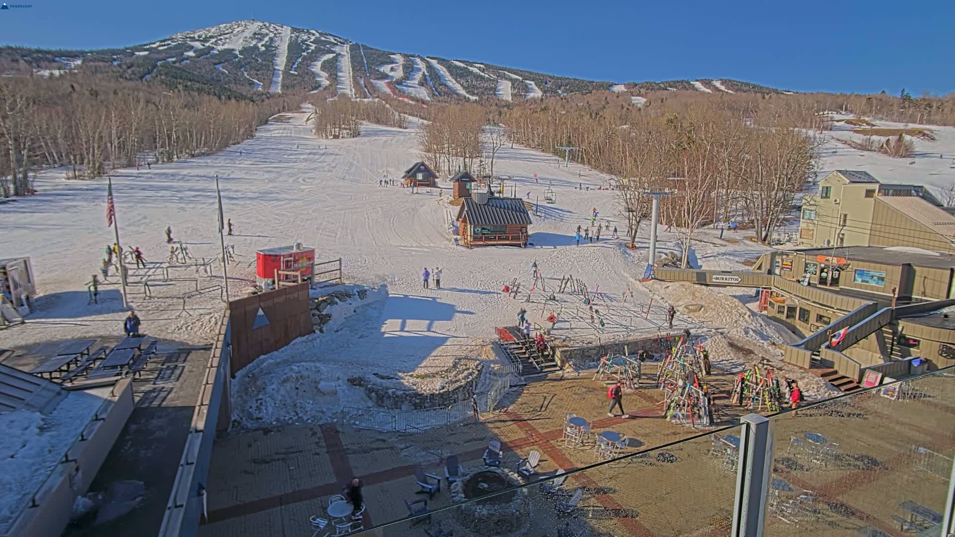 A sunny, snow-covered ski resort with several skiers and snowboarders visible on the slopes and in the base area, along with buildings and outdoor seating.