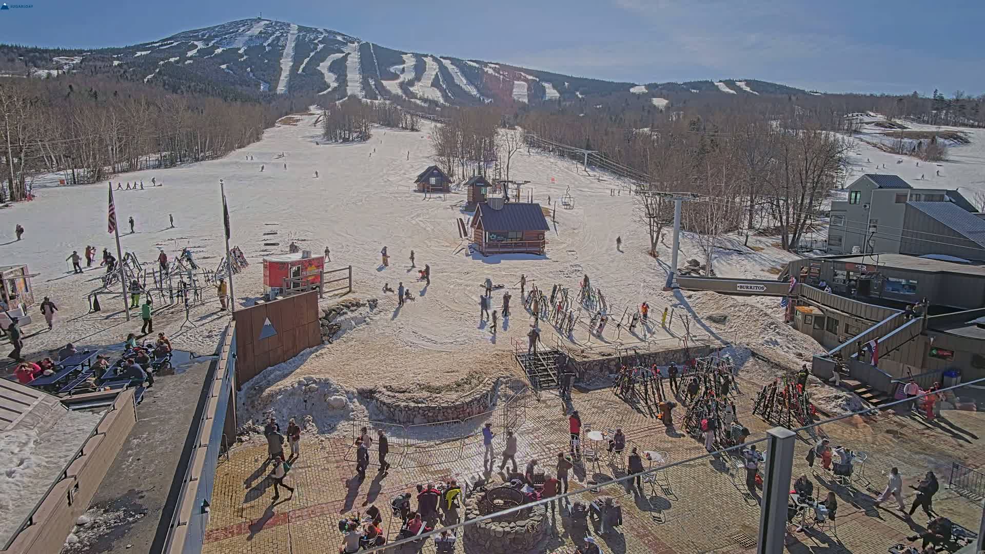 A sunny day at a ski resort shows snow-covered slopes, skiers and snowboarders, and people gathered in a patio area near ski equipment and buildings.