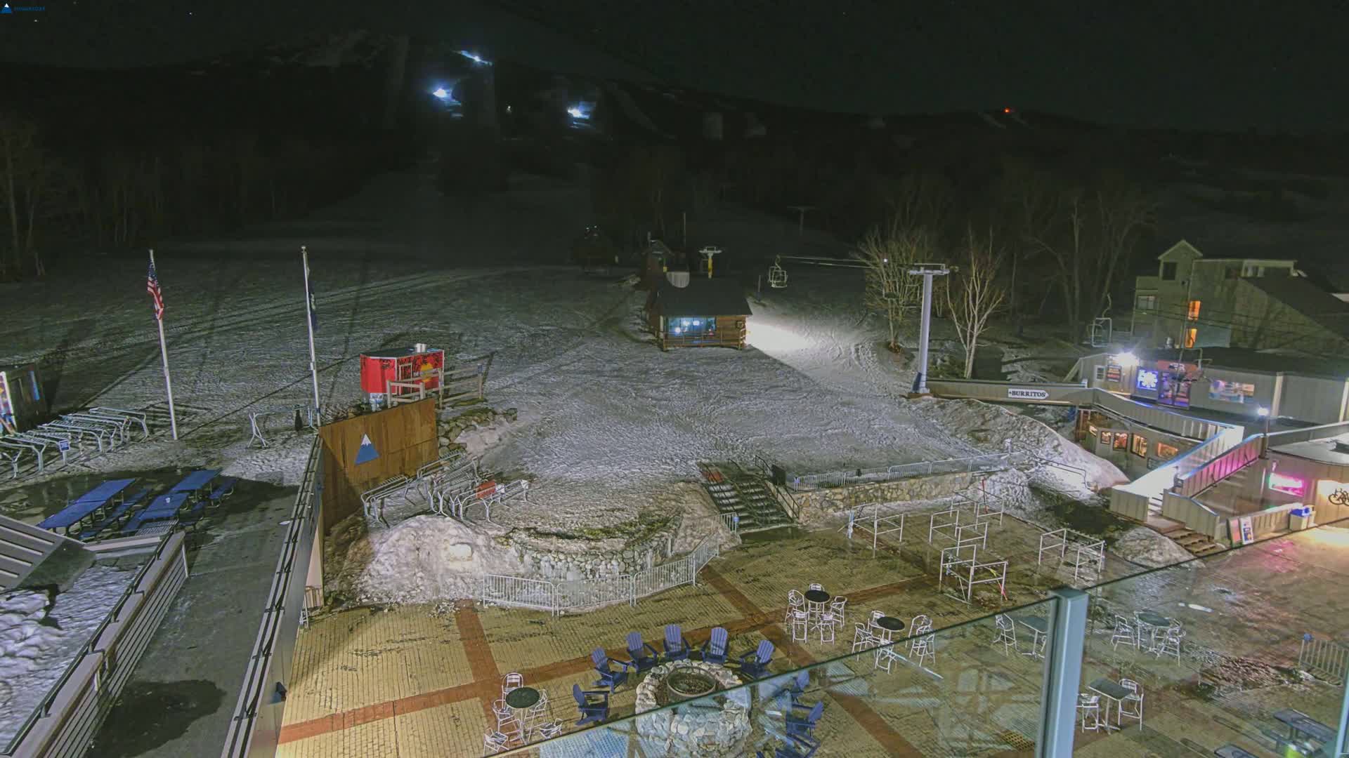 A snow-covered ski resort at night, illuminated by artificial lights, features a patio with seating around a fire pit, a ski lift, and several buildings.