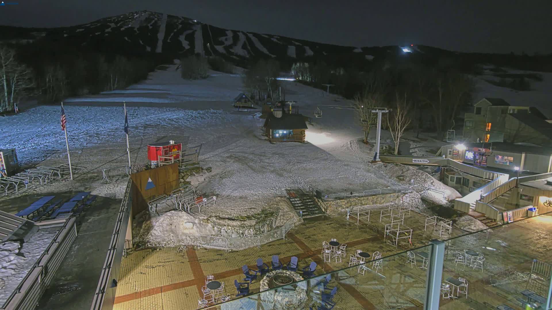 A snow-covered ski resort at night, illuminated by artificial lights, showing a base area with outdoor seating and a small building, with a mountain in the background.