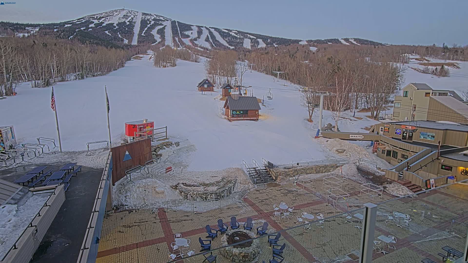 A snow-covered ski resort base area with several small buildings, outdoor seating areas, and a ski mountain in the background under a clear, twilight sky.