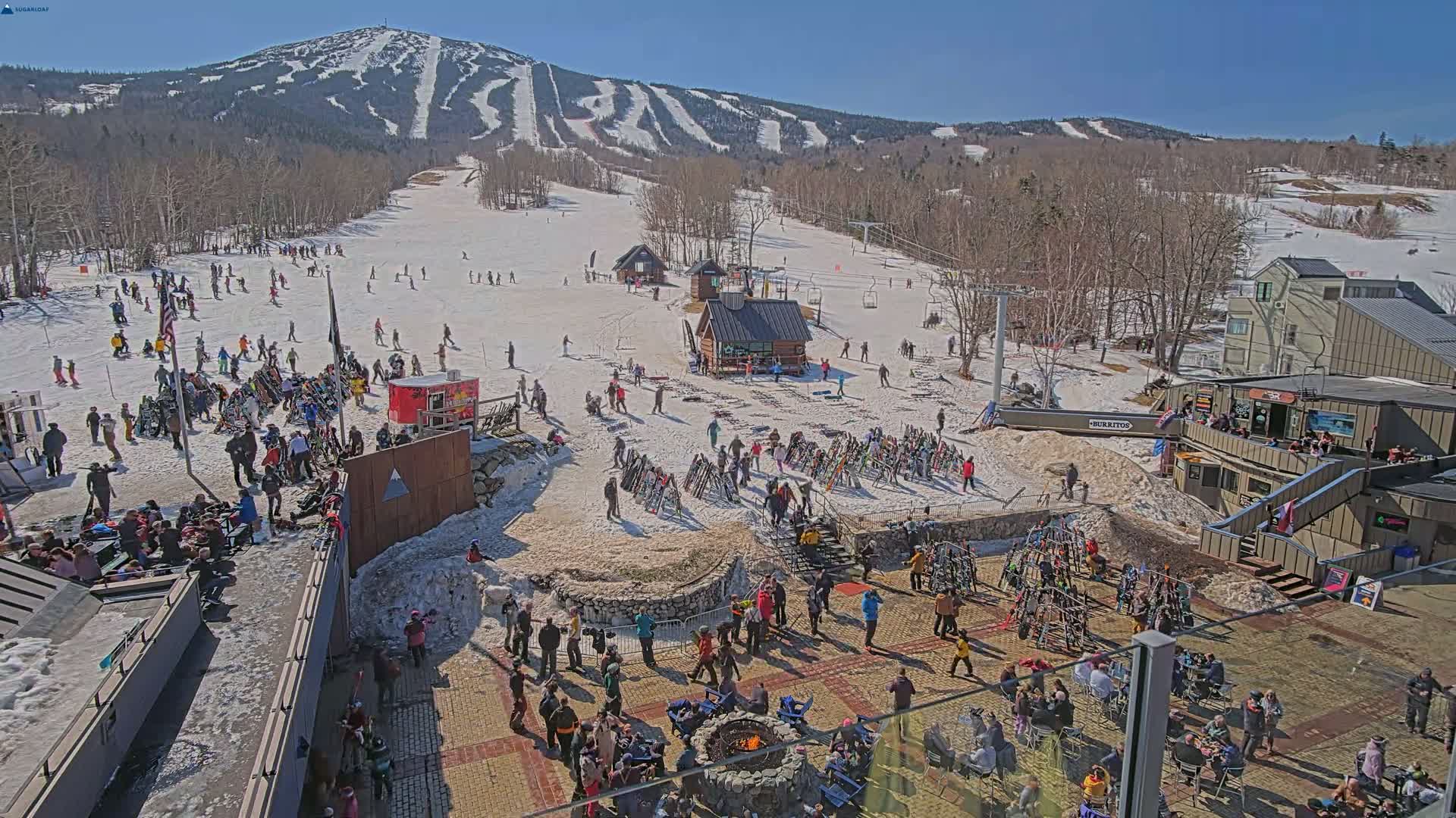 A sunny day at a ski resort shows numerous skiers and snowboarders on the slopes and gathered around a fire pit in a plaza at the base of a snow-covered mountain.