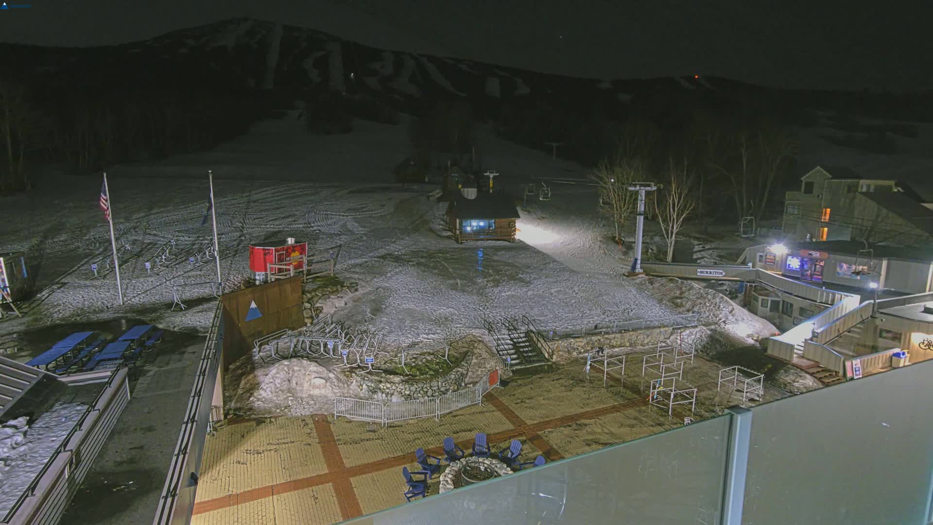 A nighttime view of a snow-covered ski resort base area with a fire pit, seating, and ski lifts, under a clear, dark sky.