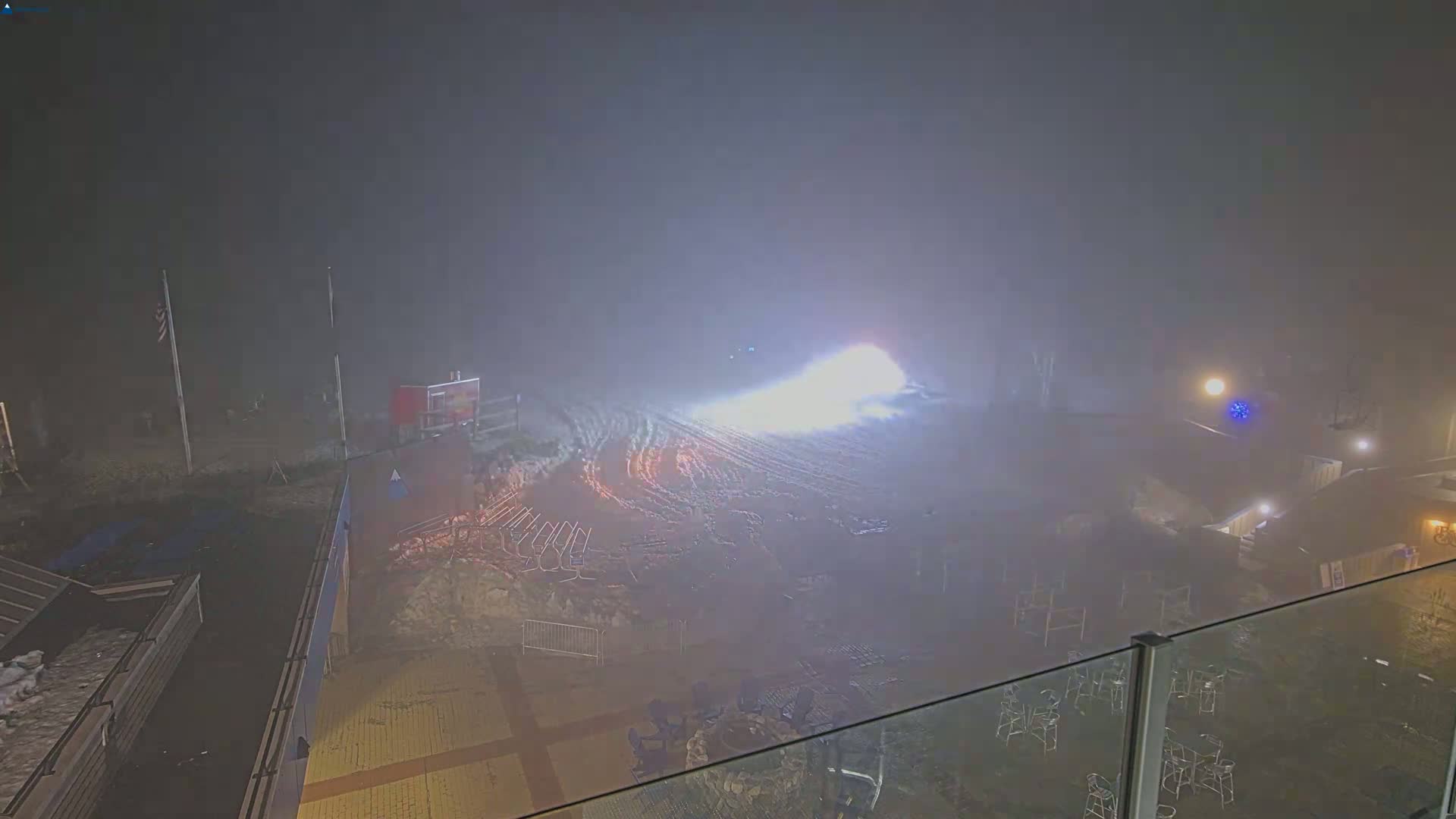 A snowy, foggy night scene shows a brightly lit ski slope with outdoor seating areas and buildings in the foreground.