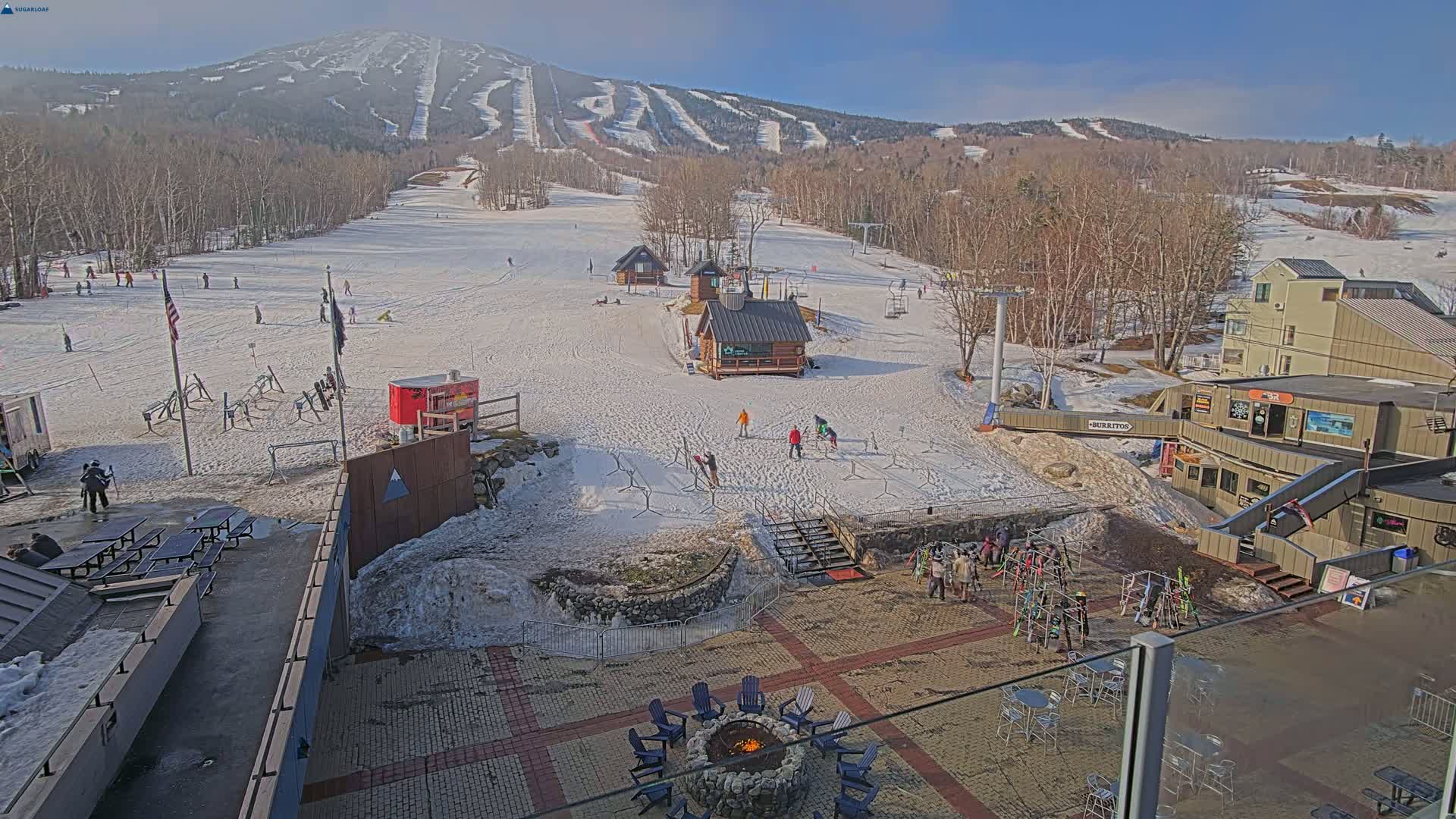 A snow-covered ski resort with several buildings, skiers and snowboarders, and a fire pit in a patio area on a sunny day.