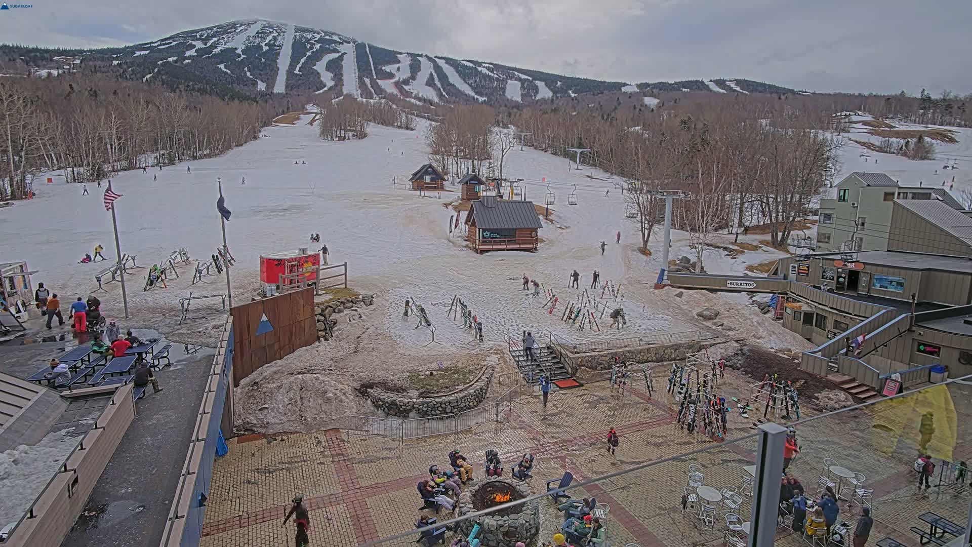 A snowy ski resort base area with people gathered around a fire pit and others skiing on the slopes under an overcast sky.