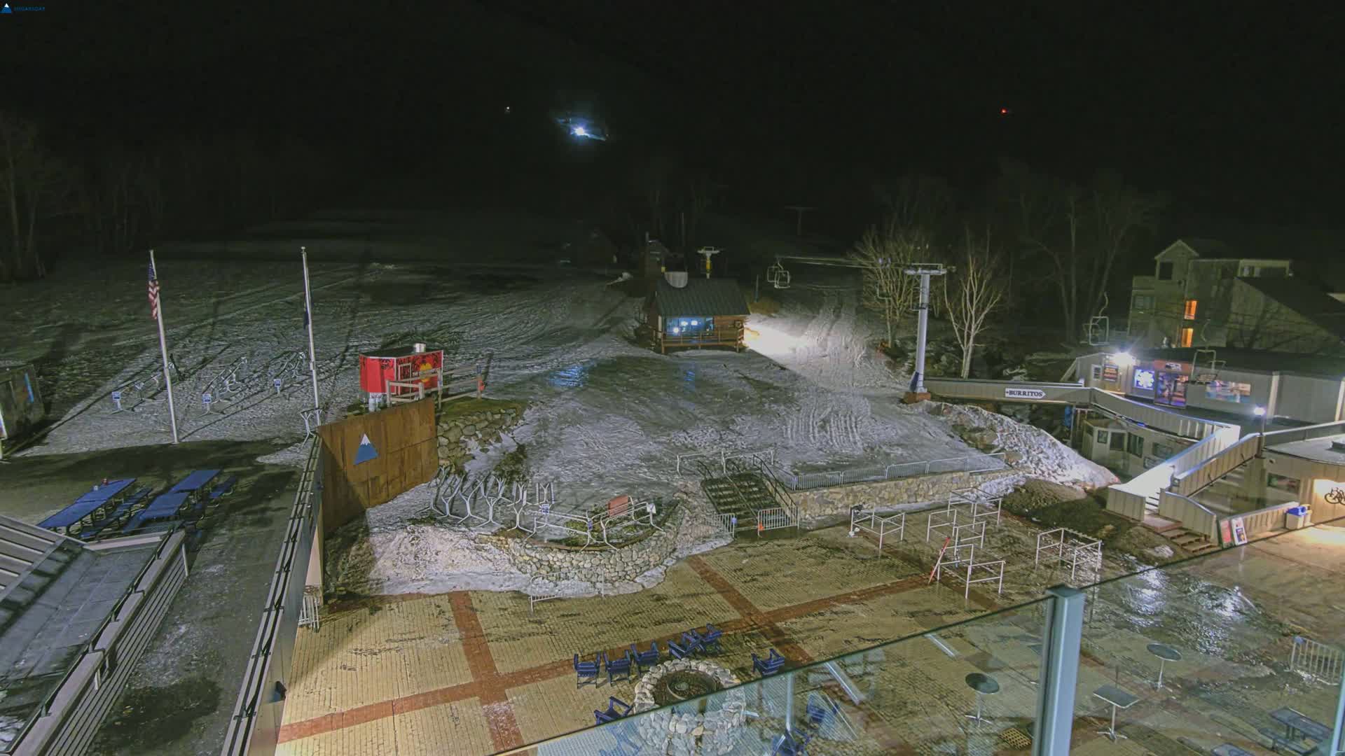 A snow-covered ski area at night, illuminated by artificial lights, shows a base lodge, ski lift, and outdoor seating areas.