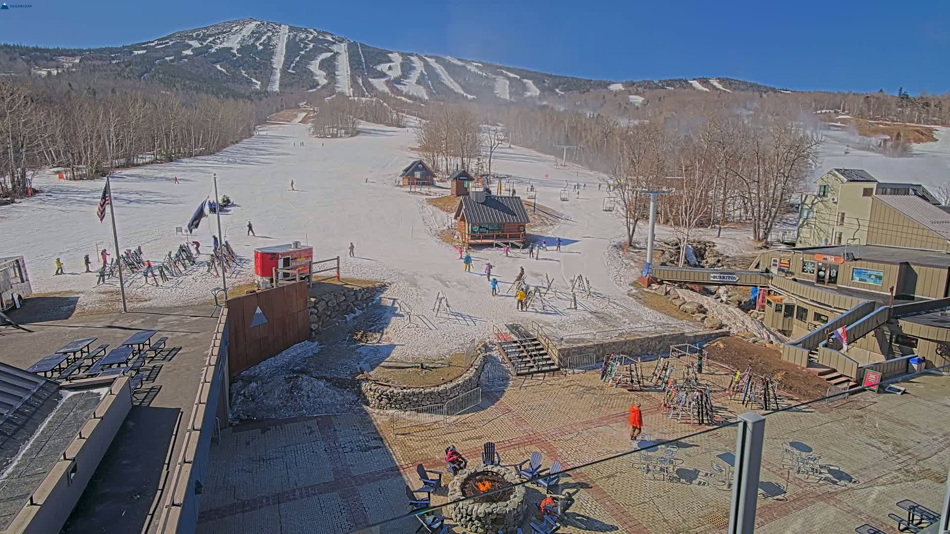 A sunny view of a snow-covered ski resort with people scattered throughout the base area, a fire pit, and a mountain in the background.