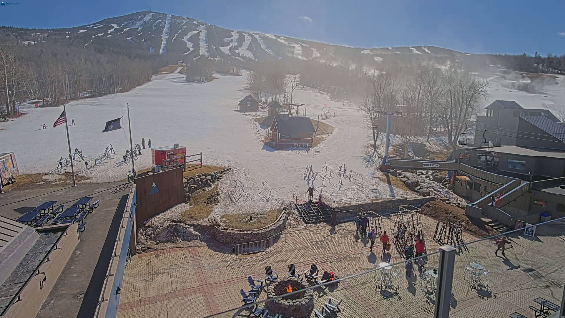 A sunny day at a ski resort shows a snow-covered mountain in the background, with a paved area at the base featuring a fire pit and several people, along with buildings and snowmaking equipment.