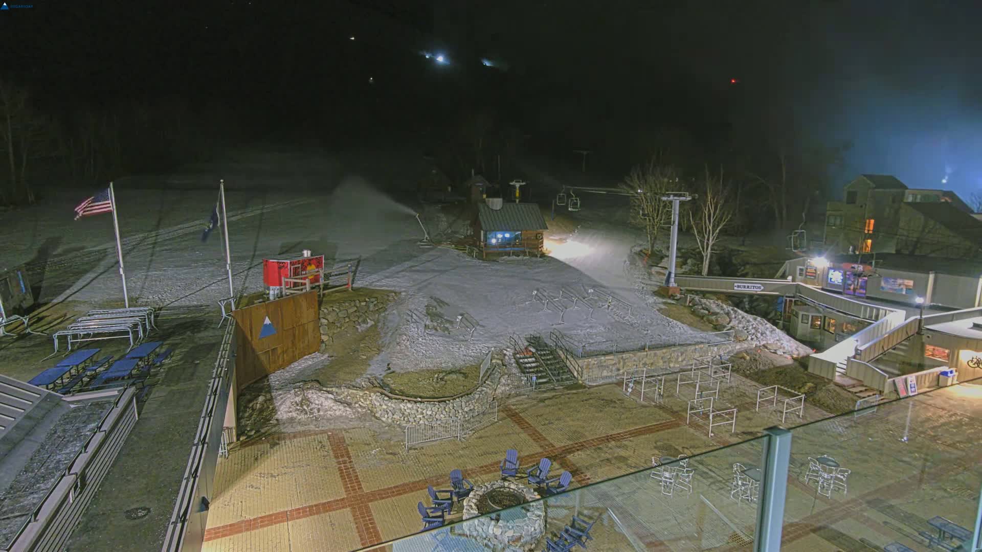 A nighttime view of a snow-covered ski slope with a snowmaking machine operating, illuminated buildings, and outdoor seating areas.